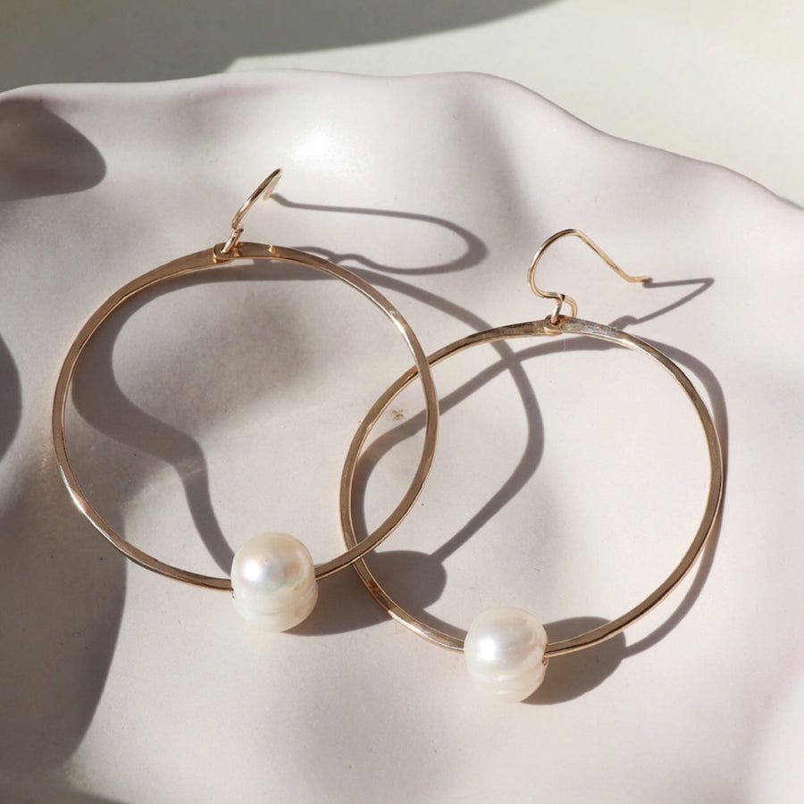 14k gold fill Pearl Hoops laid on a white plate in the sunlight. These earrings feature a hammered hoop with a pearl gemstone connecting the hoop together.