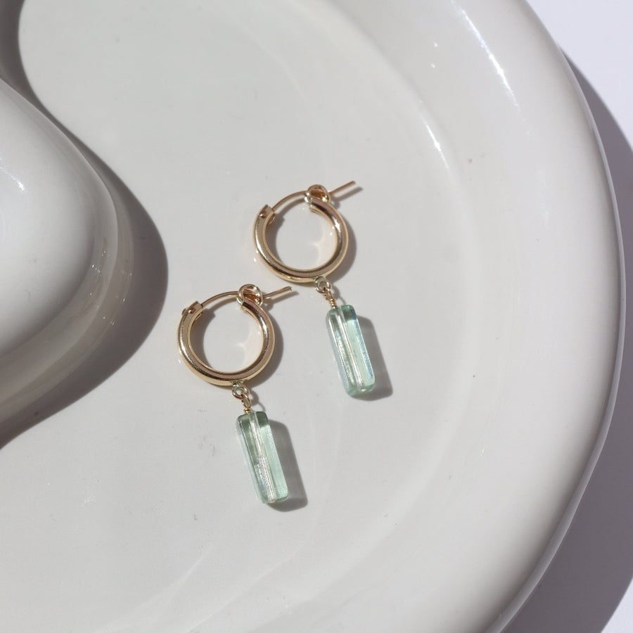 14k gold fill Flynn Hoops laid on a tan plate in the sunlight. These earrings feature a hoop like earring with a dangle of a Fluorite tube gemstone.