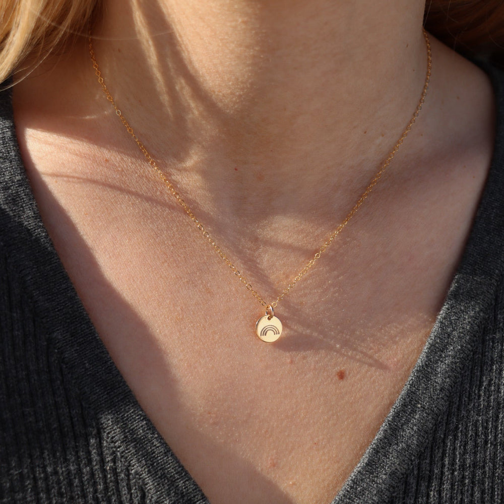 Close-up of a person wearing a delicate gold necklace with a small hand stamped rainbow pendant.