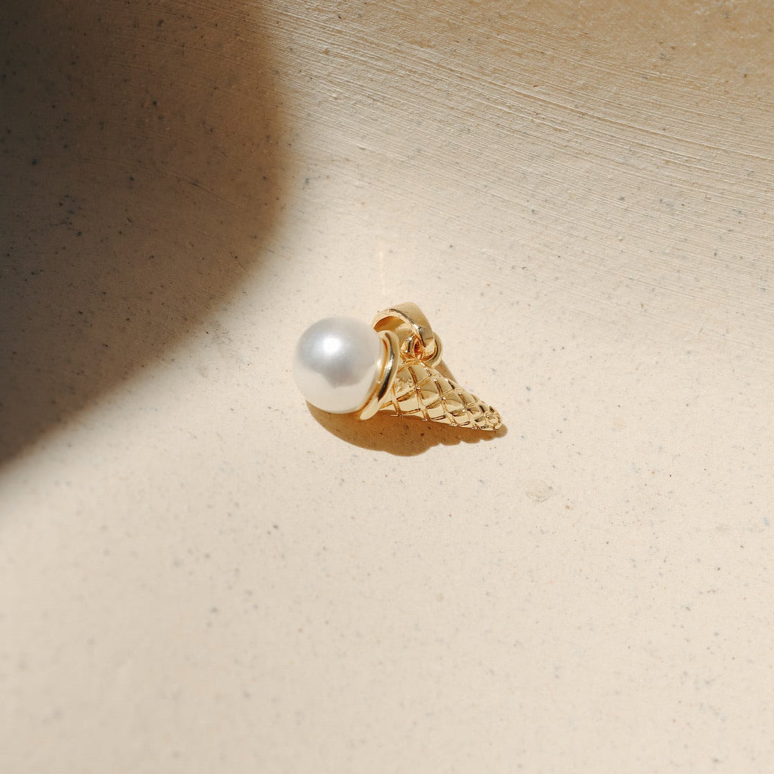 Gold ice cream cone charm featuring a lustrous round pearl scoop in a waffle textured cone, photographed on a neutral surface in warm natural light.