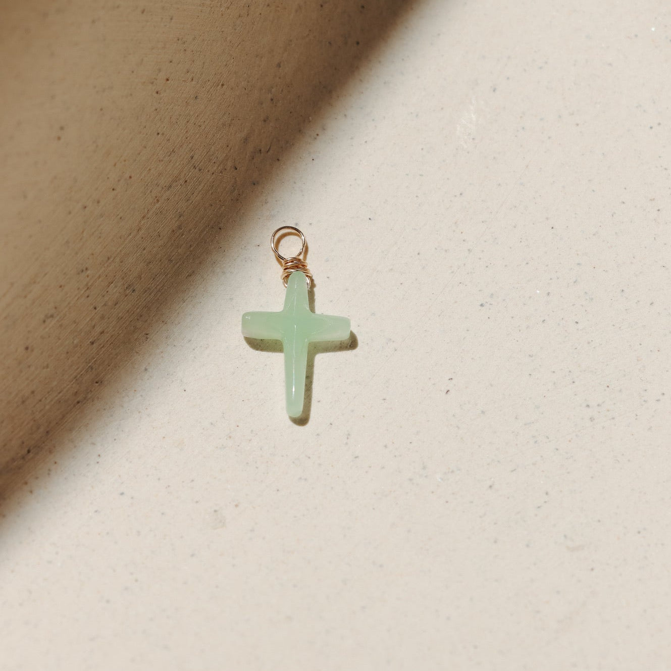 Chrysoprase cross charm featuring a soft green gemstone cross wrapped with gold wire, photographed on a warm neutral surface with soft natural light and shadows.