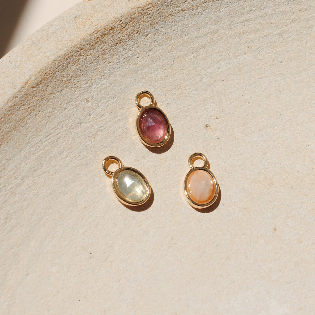 Three oval gemstone charms in pink, green, and peach tones set in gold bezels, displayed on a neutral textured surface in warm natural light.