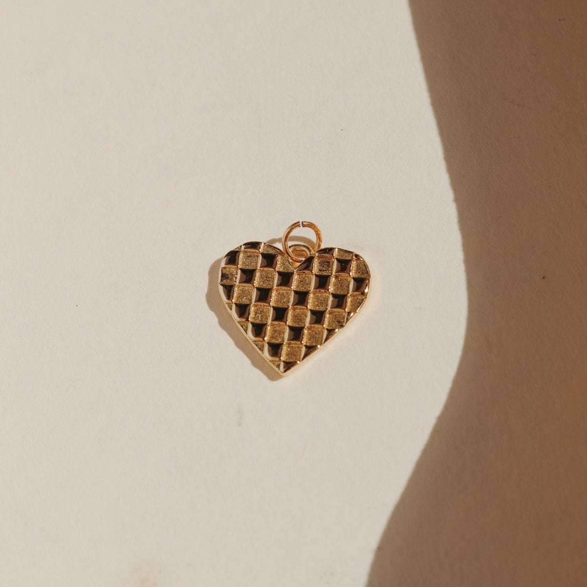 Gold plaid heart charm with a textured checkerboard pattern, photographed on a warm neutral surface with soft natural light and shadows.