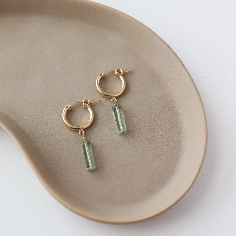 14k gold fill Flynn Hoops laid on a tan plate in the sunlight. These earrings feature a hoop like earring with a dangle of a Fluorite tube gemstone.