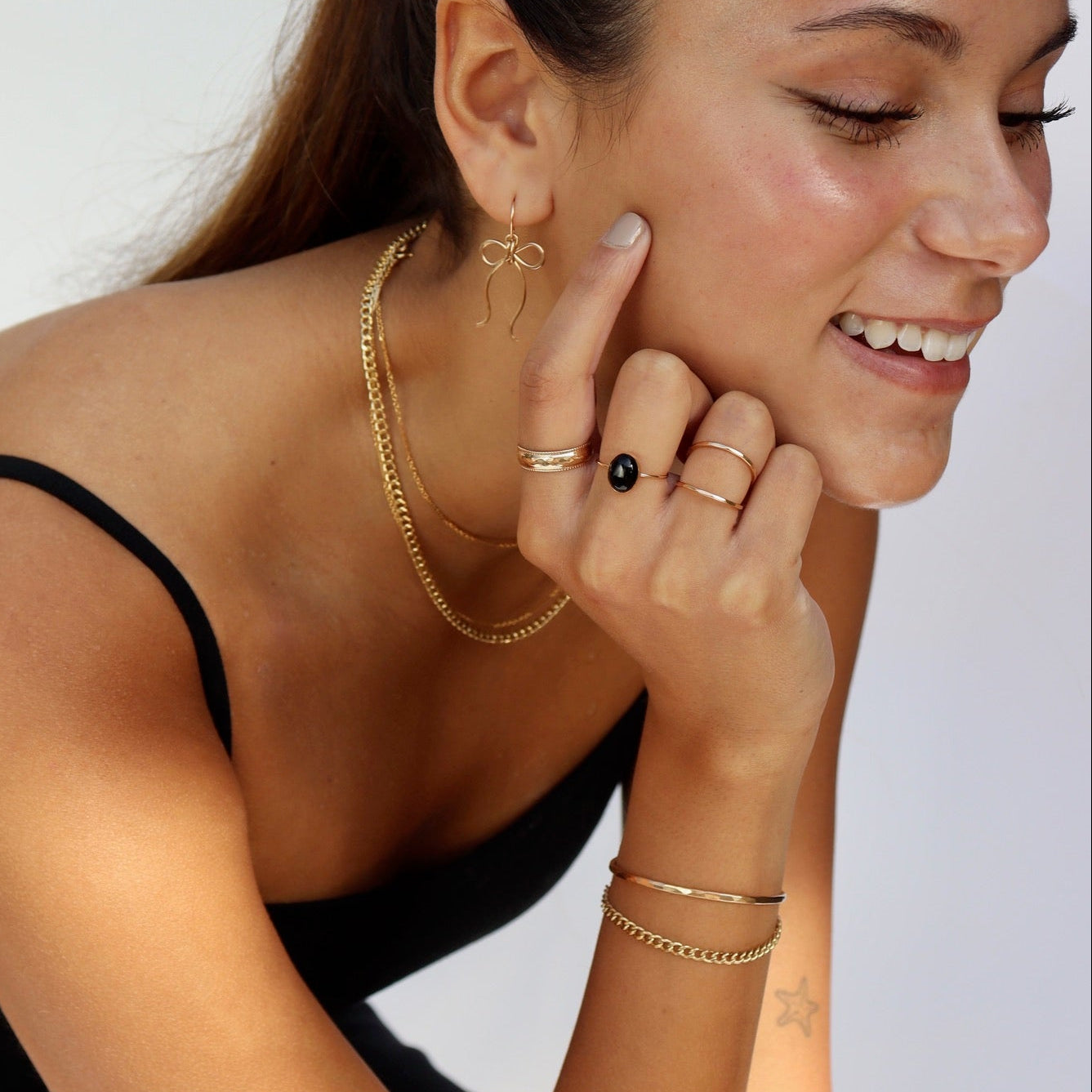 Side profile of a smiling dark-haired woman in a black tank top resting her face on her hand, displaying a complete gold jewelry set: delicate wire ribbon bow drop earrings, an oval black gemstone ring paired with thin gold bands, a classic layered gold chain necklace stack, and dual gold wrist cuffs.