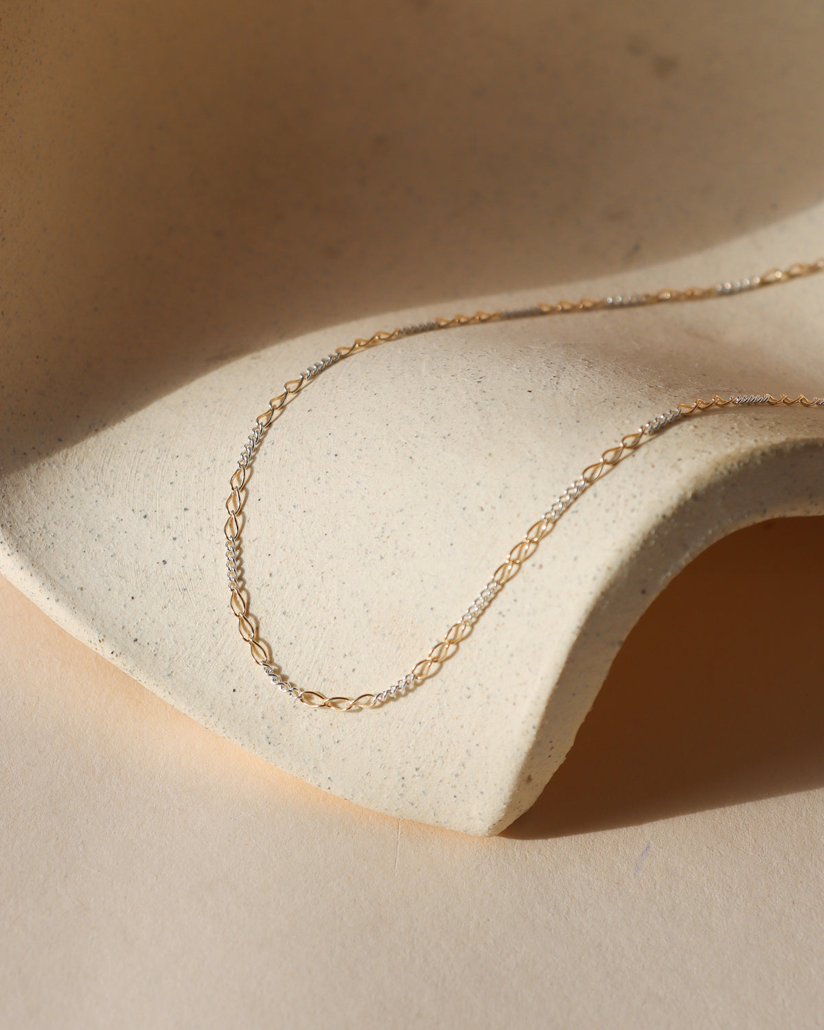 Interlude mixed metal chain necklace featuring alternating elongated 14k gold fill links and sterling silver chain links, draped over a cream ceramic surface in sunlight.