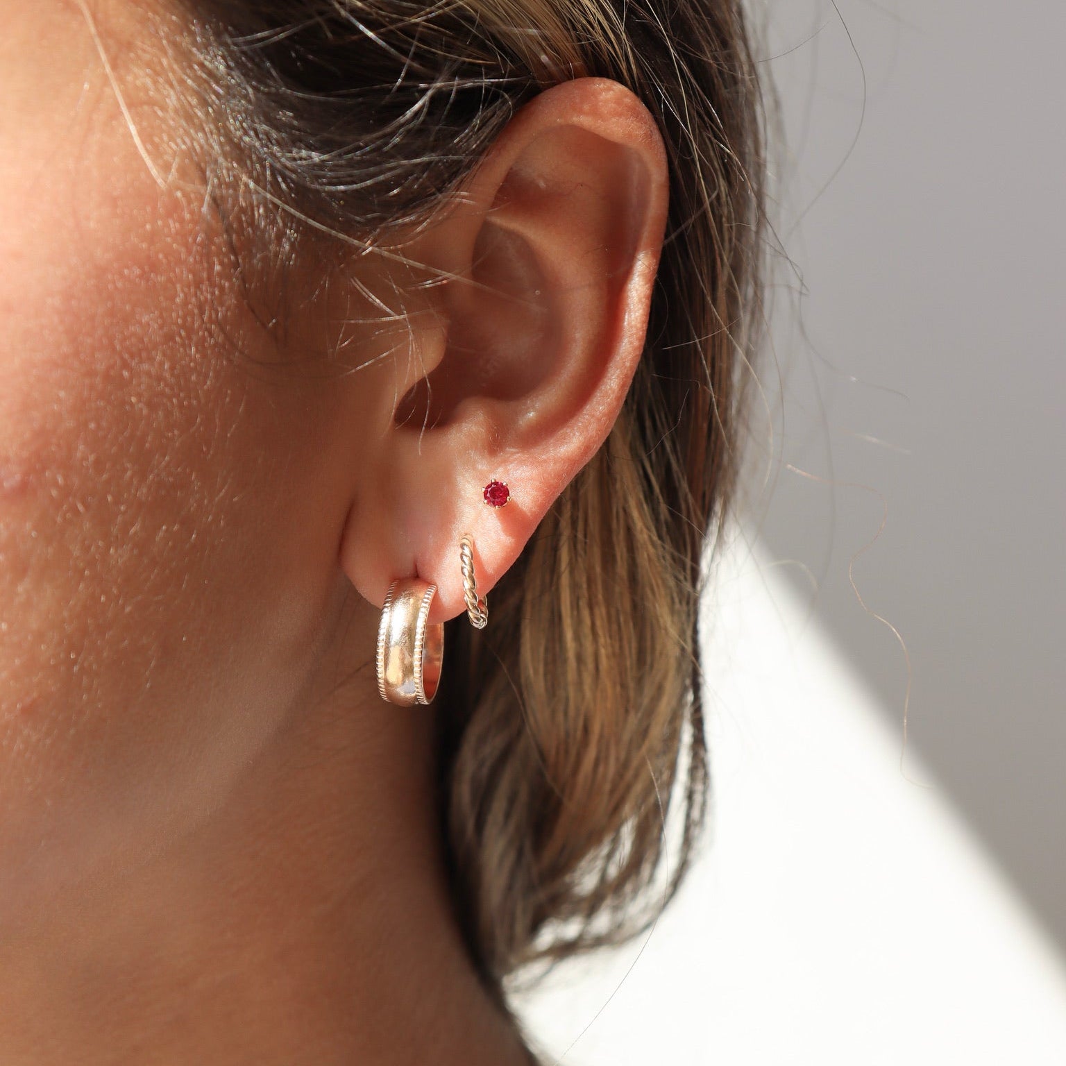 Side profile close-up showing a layered ear piercing stack. The lower earlobe features a small, chunky gold hoop with textured edges, followed by a delicate twisted gold huggie hoop, and a small round ruby-red stud higher on the lobe