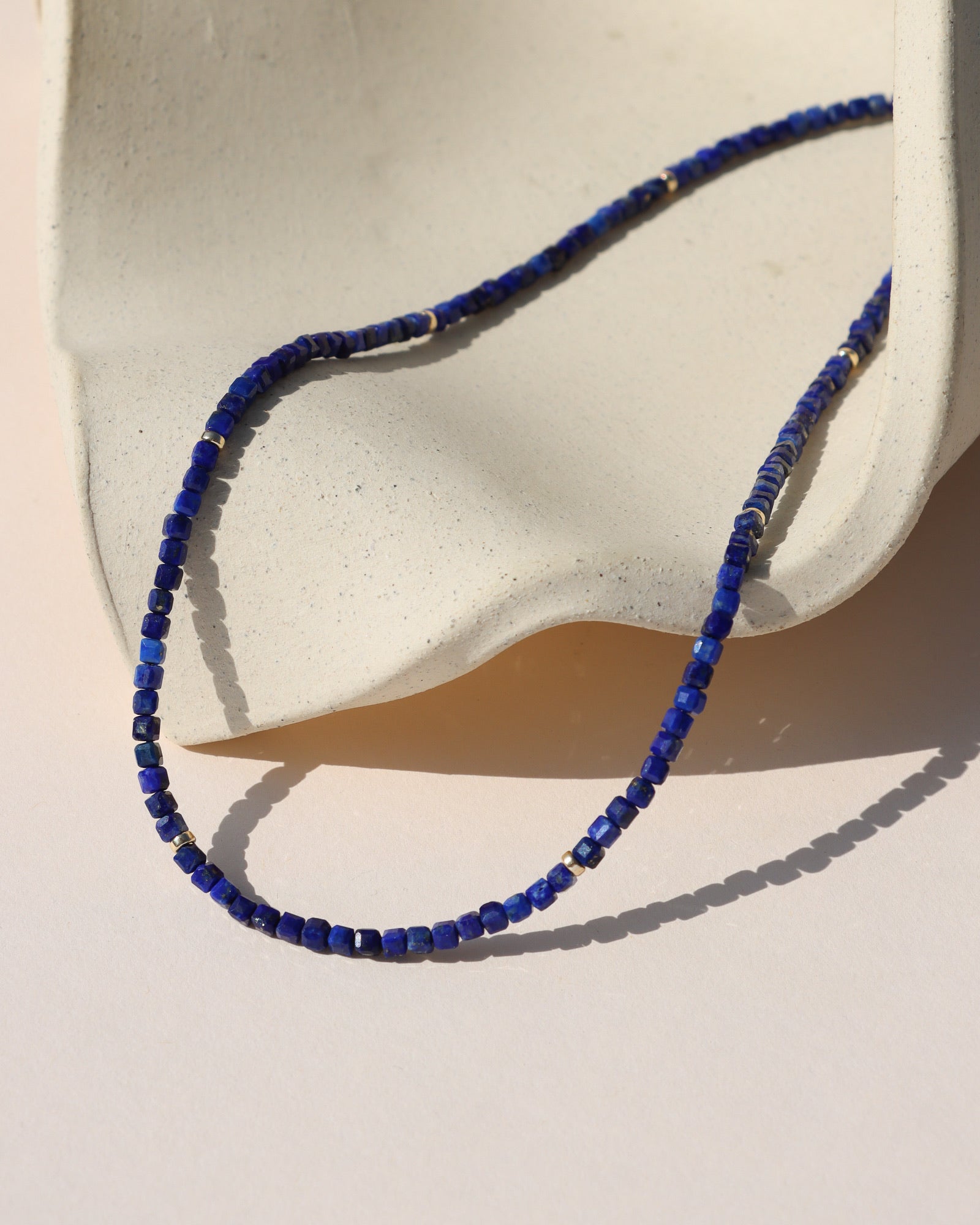 A beaded deep blue lapis necklace with small gold accent beads draped over a neutral ceramic surface.
