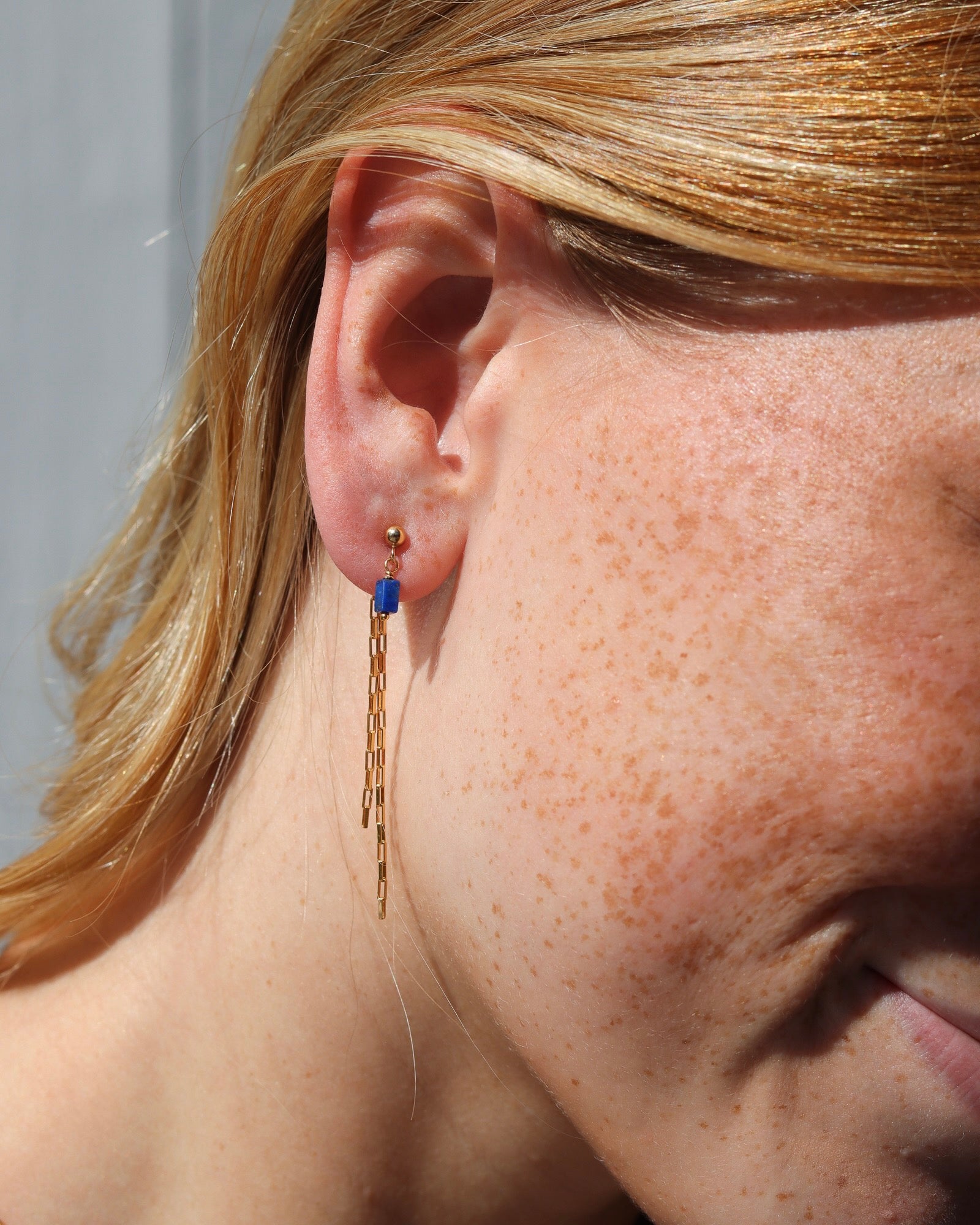Close-up of a person wearing a gold stud earring with a small blue stone dangle and long, cascading gold chain fringe.