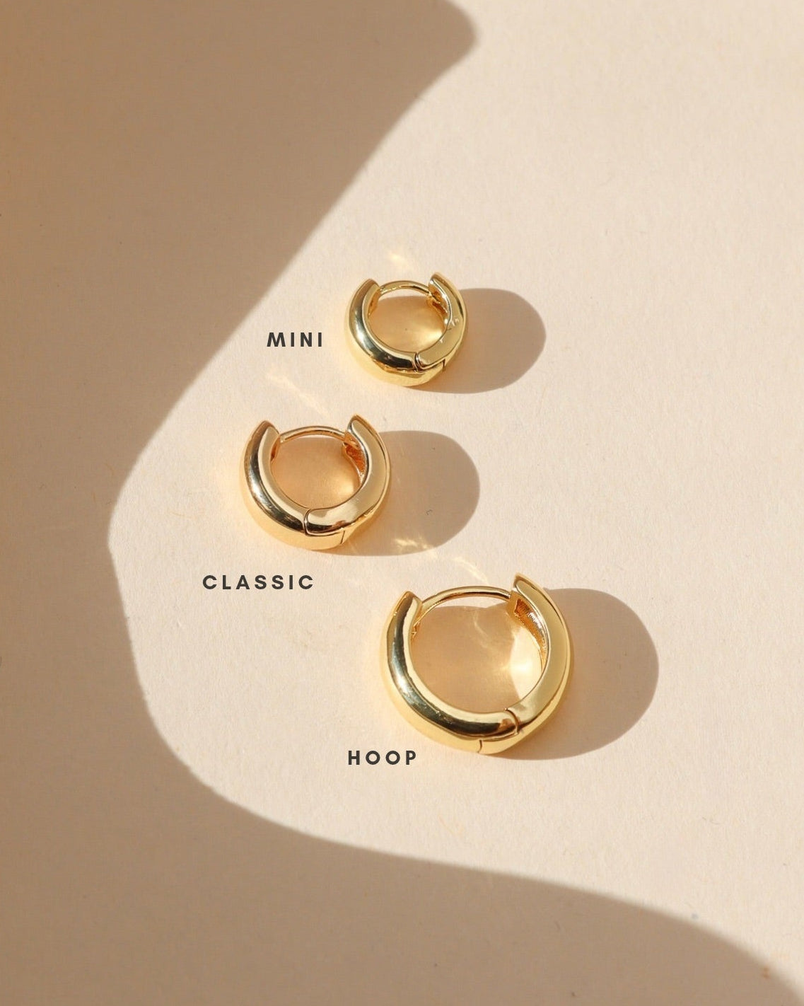 Three polished gold Luxe hoop earrings displayed in Mini, Classic, and Hoop sizes on a cream background in warm natural sunlight, showing the differences in size and rounded design.