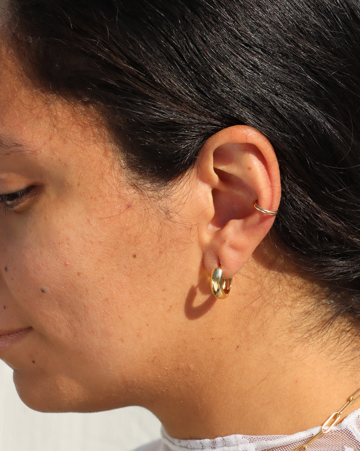 Close-up of a model wearing a chunky polished gold hoop earring with a slim gold ear cuff on the upper cartilage, styled in warm natural sunlight.