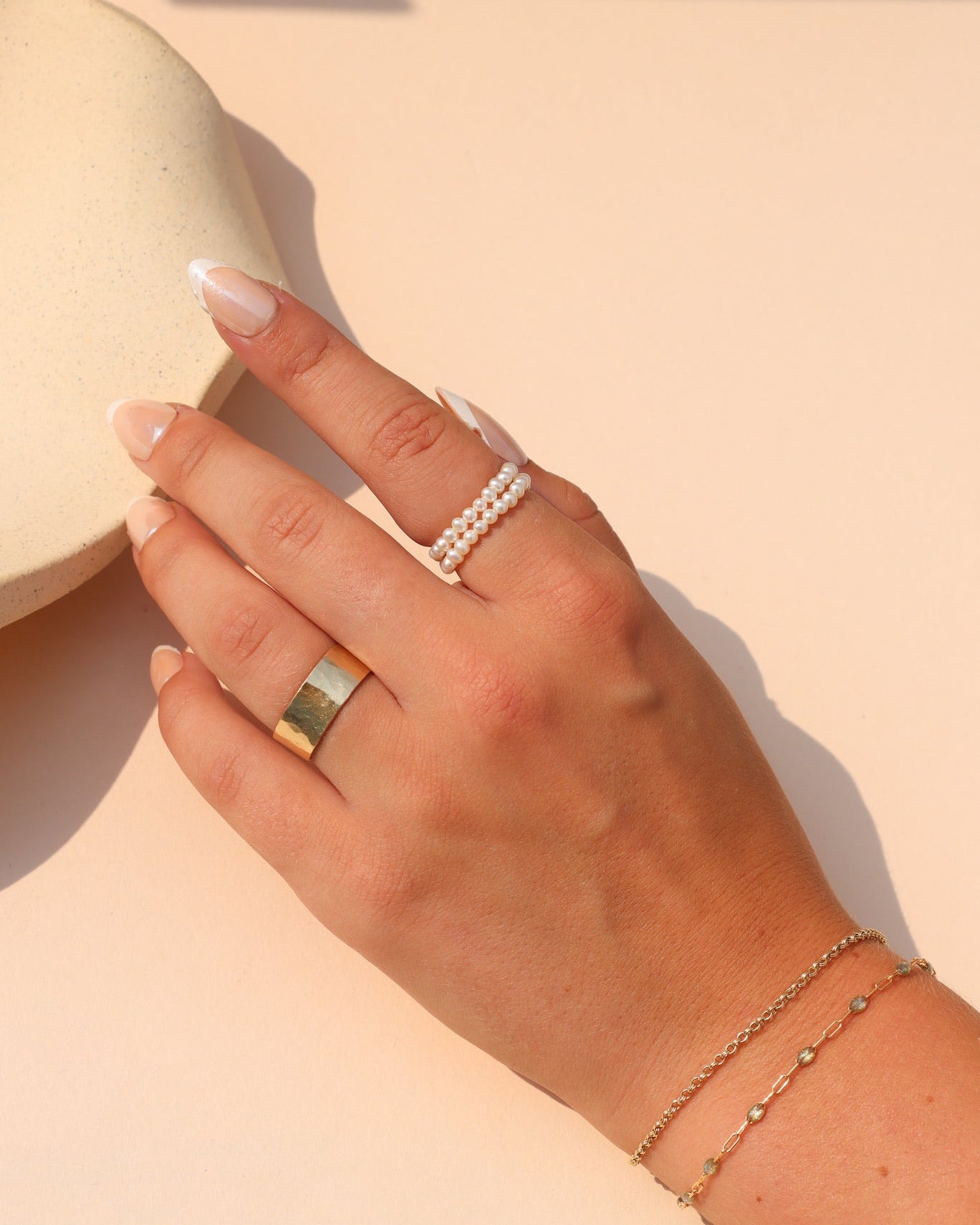 Delicate double-row pearl ring styled with a gold statement ring and layered gold bracelets in warm sunlight.