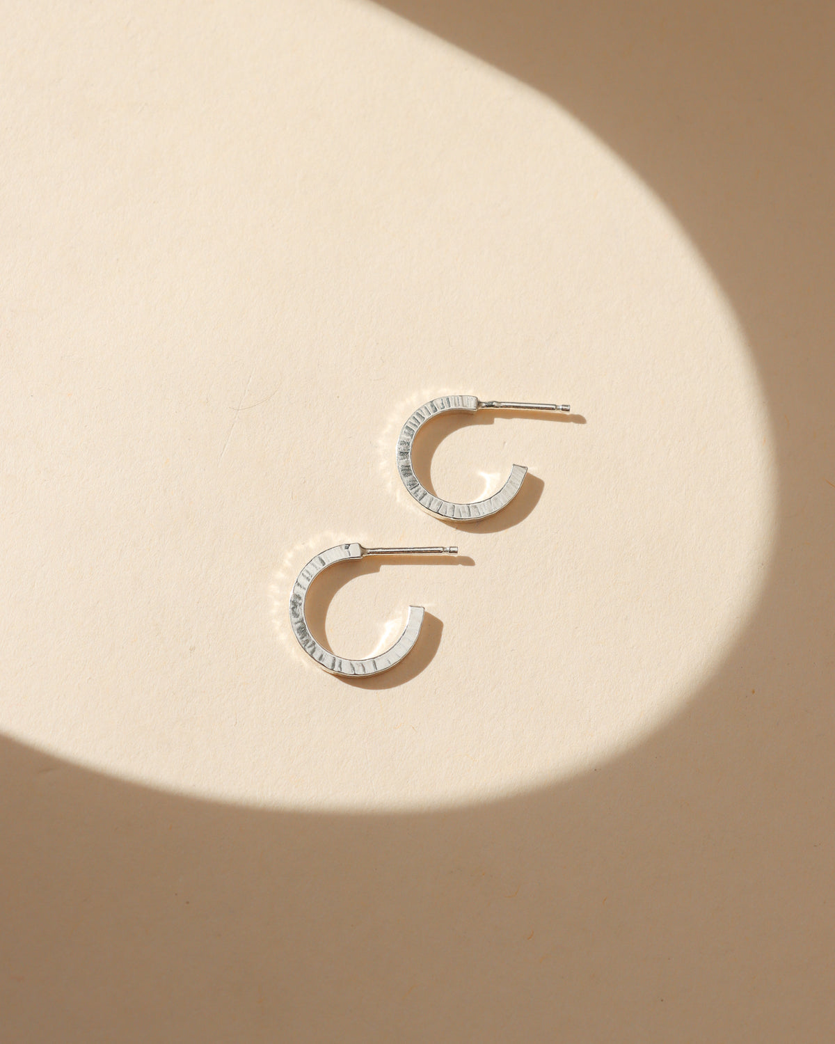 Small sterling silver huggie hoop earrings with a textured, hammered finish resting on a cream background with soft shadows.