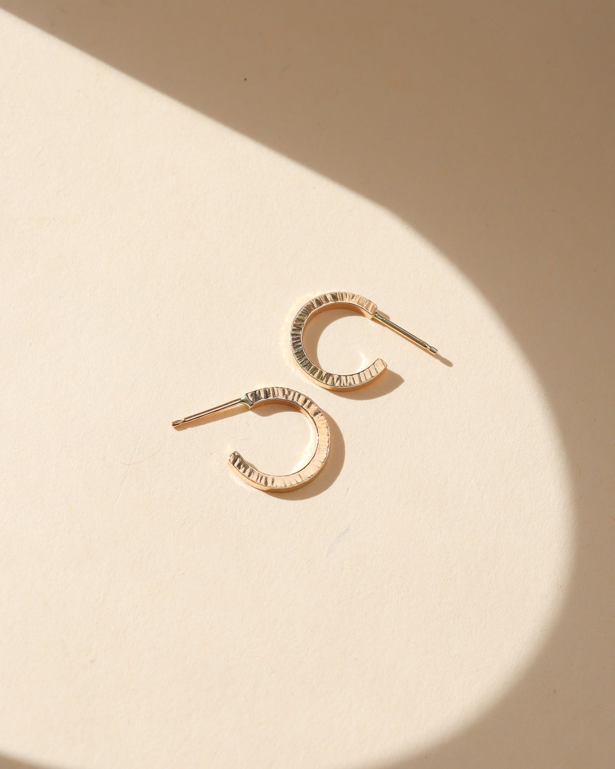 Small gold huggie hoop earrings with a textured, hammered finish displayed on a cream background in warm natural sunlight.