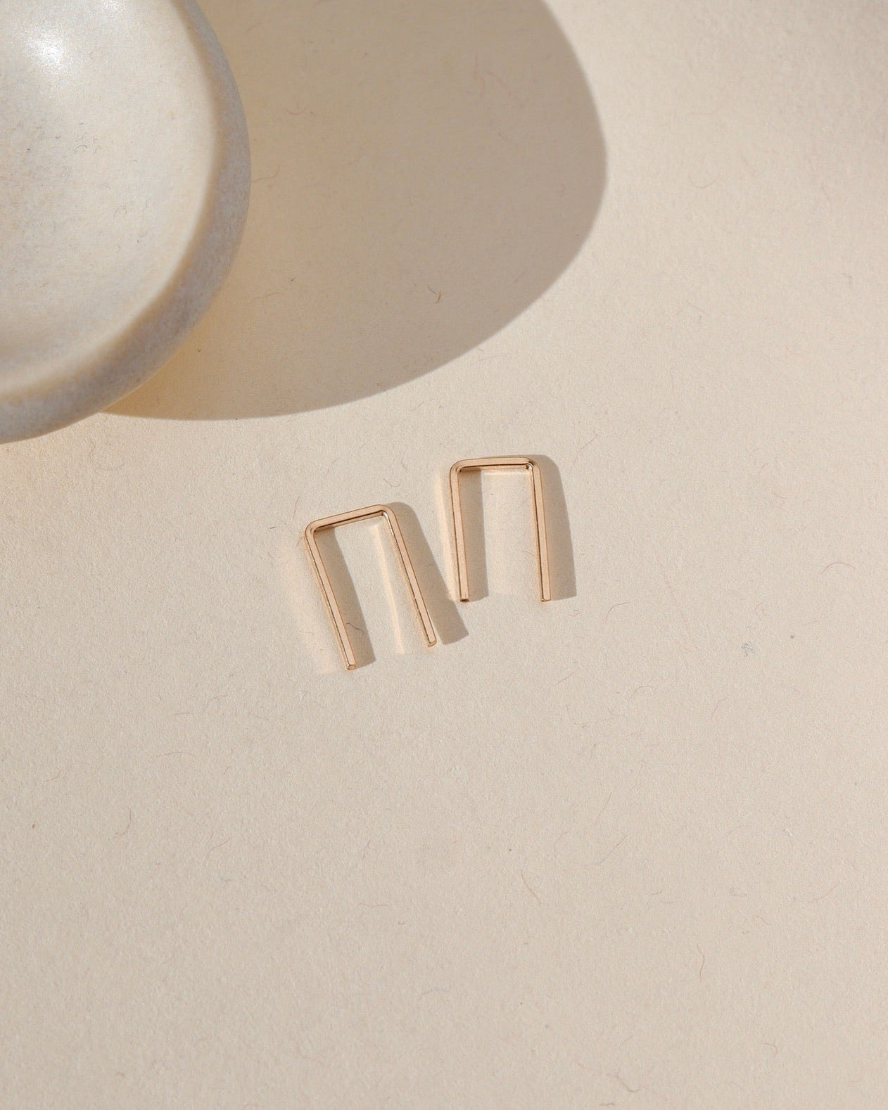 14k gold fill mini staple shaped earrings laid by small dish in the sunlight