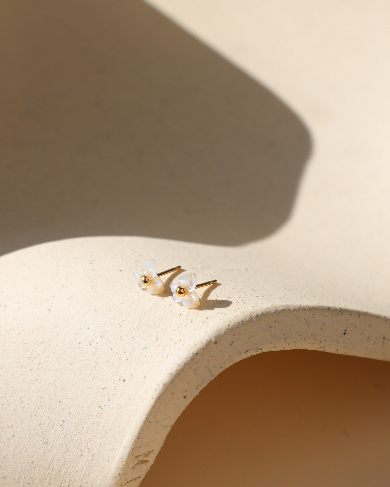 Pair of mother-of-pearl flower stud earrings with polished 14k gold fill centers resting on a textured cream ceramic display in warm natural light.