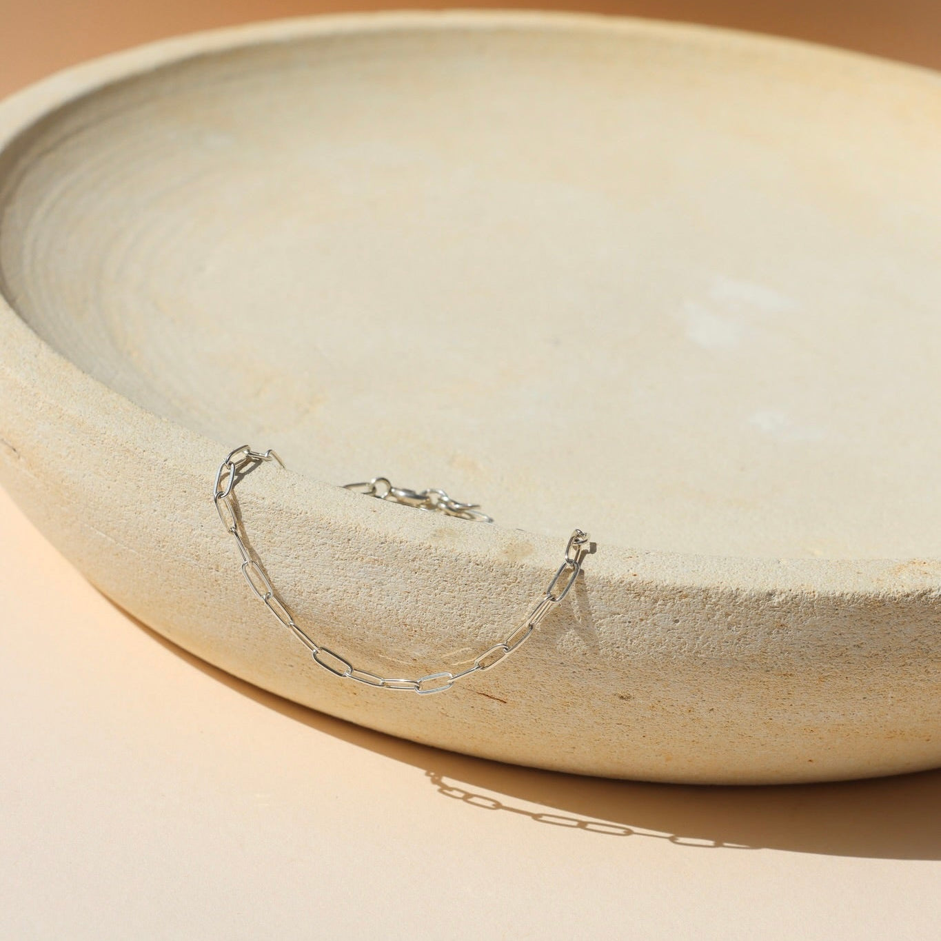 A delicate silver paperclip chain bracelet draped over the rim of a textured beige ceramic plate on a warm beige surface.