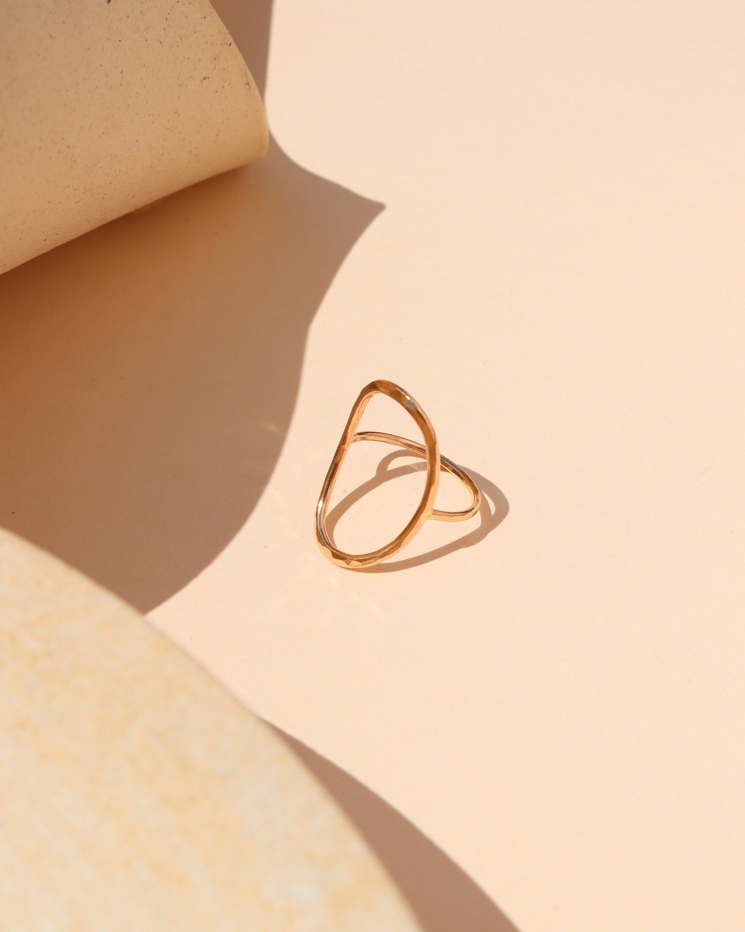 Minimalist gold ring with an open oval outline design displayed on a peach-beige surface, surrounded by curved neutral props and dramatic warm sunlight shadows.