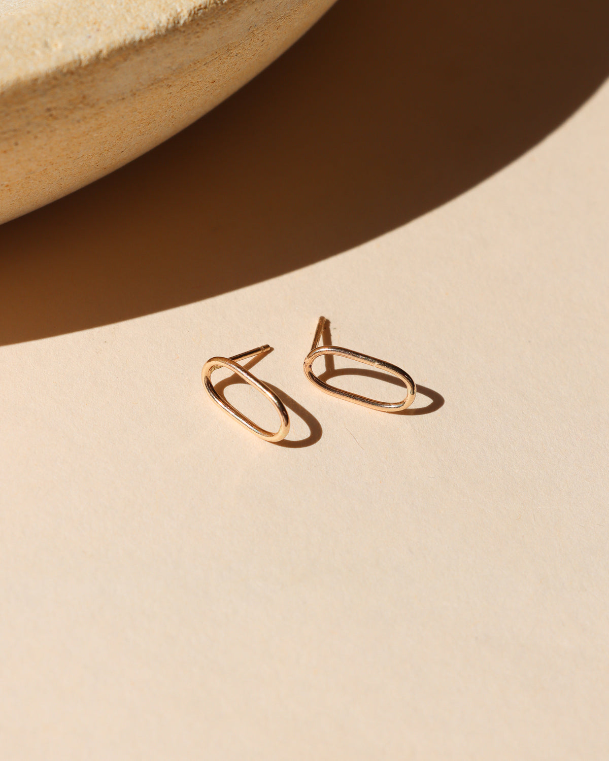 Gold-filled open oval stud earrings displayed on a neutral surface in warm sunlight.