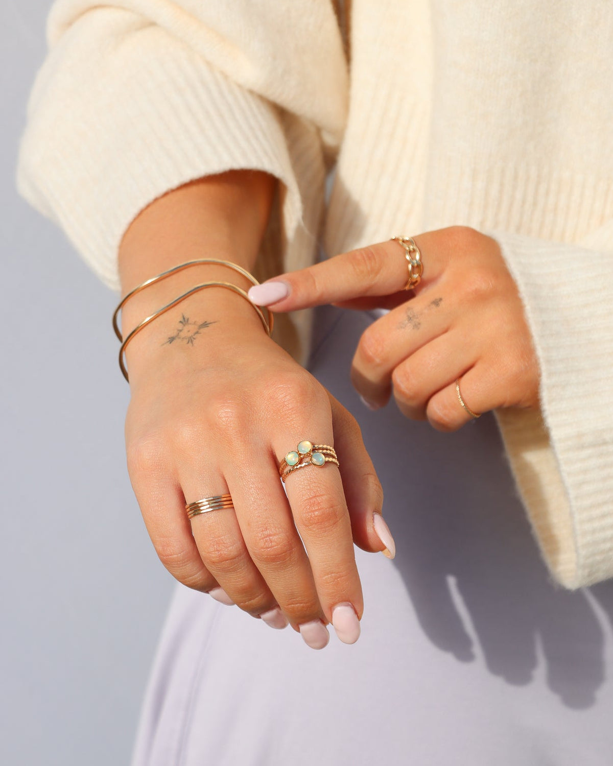 Model wearing a delicate gold opal ring, layered gold necklaces, a stacked gold band ring, photographed in warm natural sunlight.