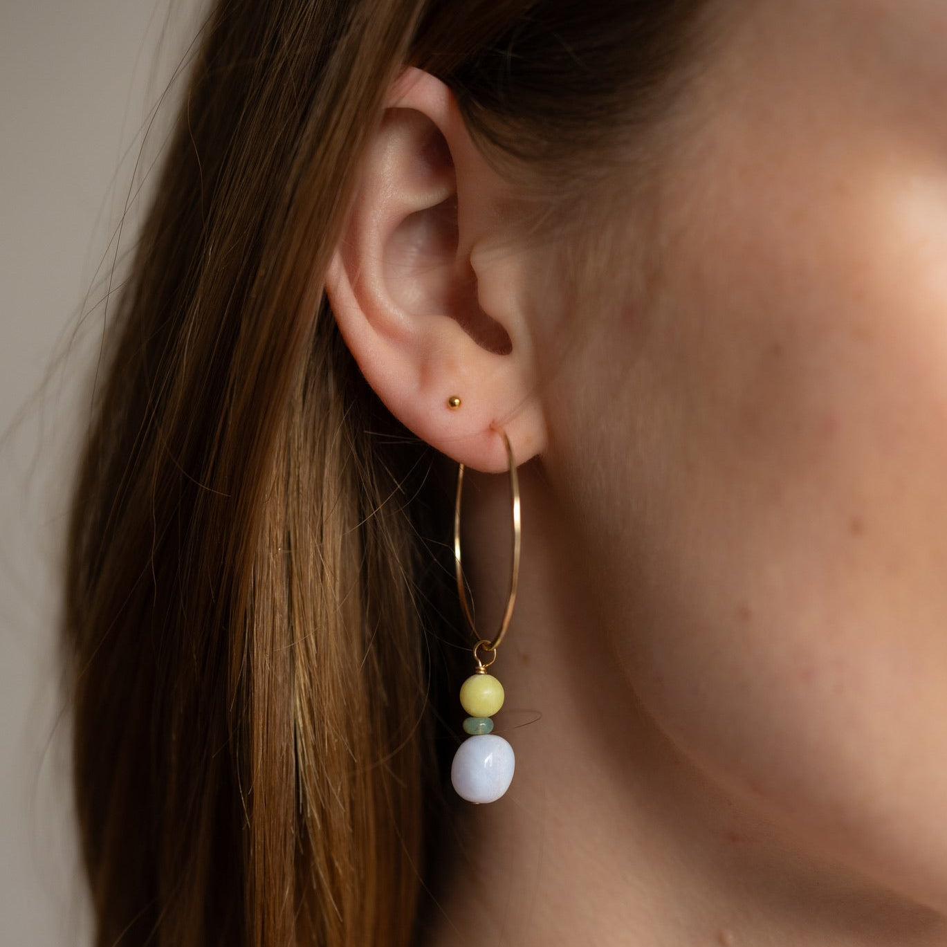 Model wearing 14k gold fill hoop earrings with dangling natural gemstone beads in soft yellow, mint green, and light blue. Minimal statement earrings styled with layered gold necklaces.