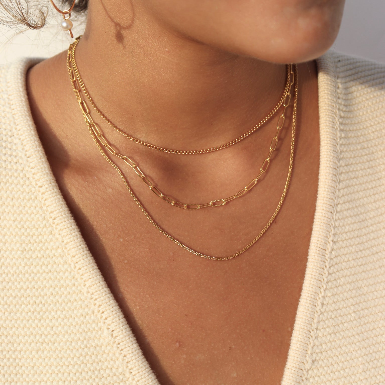 Close-up shot of a woman's neckline in soft natural lighting, showcasing a three-tier gold necklace stack: a top delicate curb chain, a middle paperclip chain, and a bottom fine woven chain, styled over a cream V-neck knit top and paired with a thin gold hoop earring featuring a dangling pearl drop