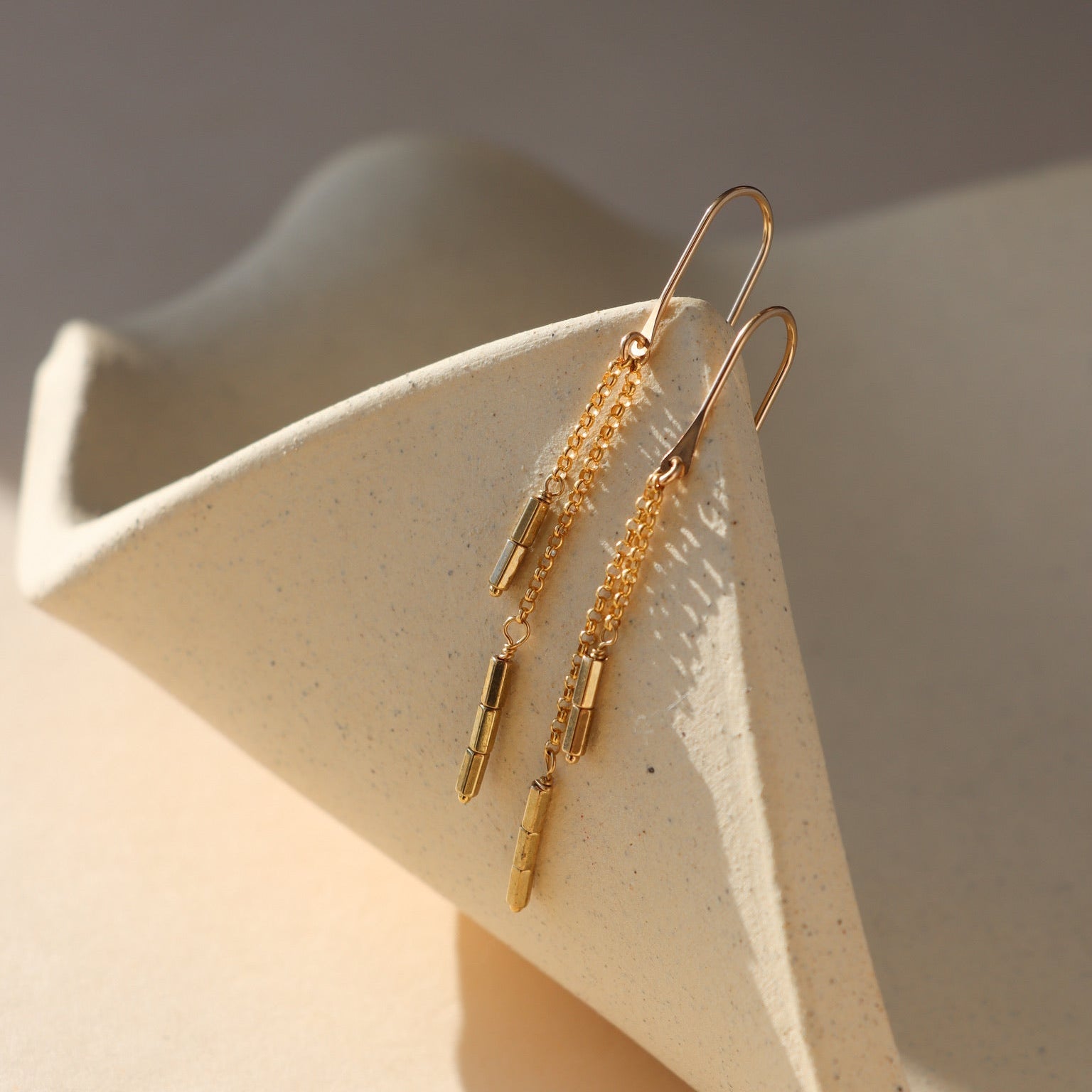 Remy chain drop earrings in gold with delicate chains and elongated accents, styled on a neutral sculptural surface.