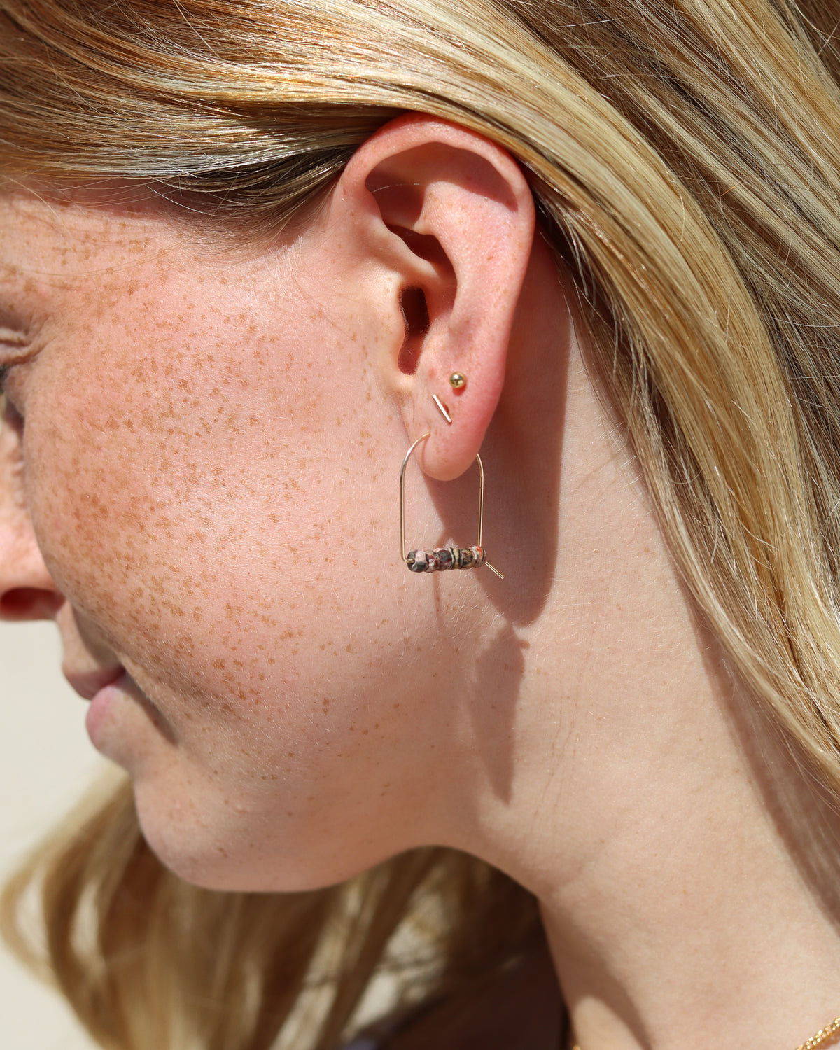 Gold arch earring with a row of small multicolored stone beads, worn alongside a gold ball stud and bar stud in warm sunlight.