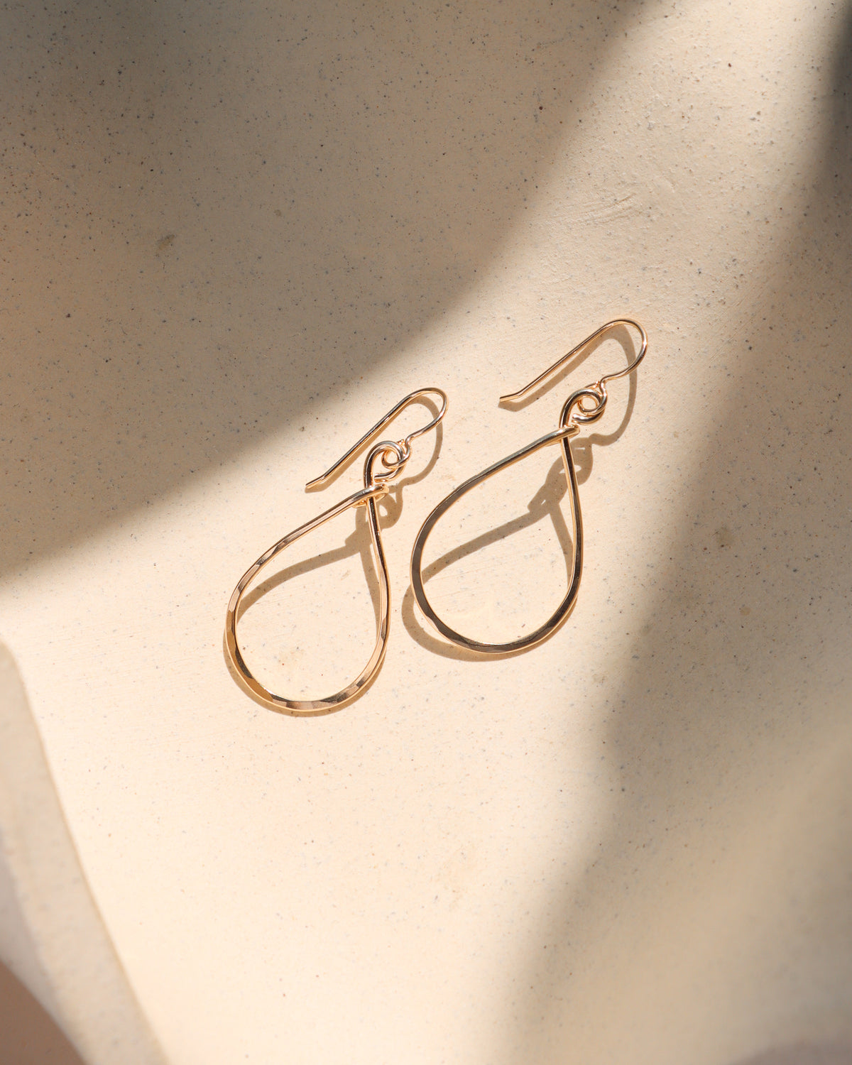 A pair of gold-toned teardrop dangle earrings with a polished finish, displayed on a textured beige surface with soft diagonal shadows.