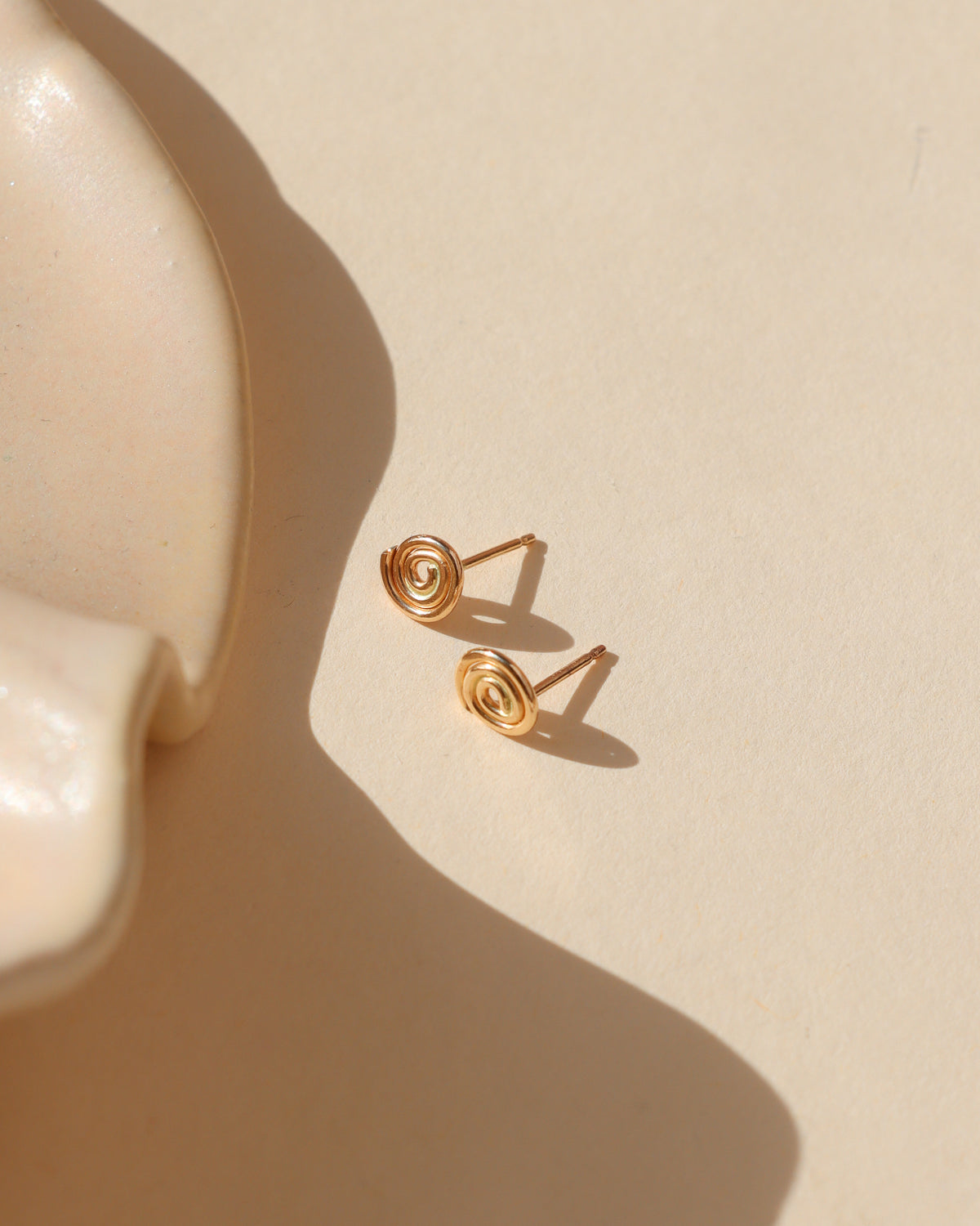 Gold spiral stud earrings featuring a delicate circular swirl design resting on a cream background with warm sunlight and soft shadows.