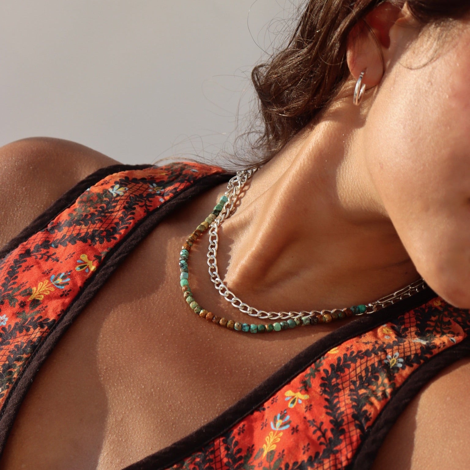 Close-up of a dark-haired woman in a patterned orange top wearing small silver hoop earrings and two layered necklaces: a chunky silver link chain paired with a bead strand of turquoise and earthy brown stones in natural sunlight.