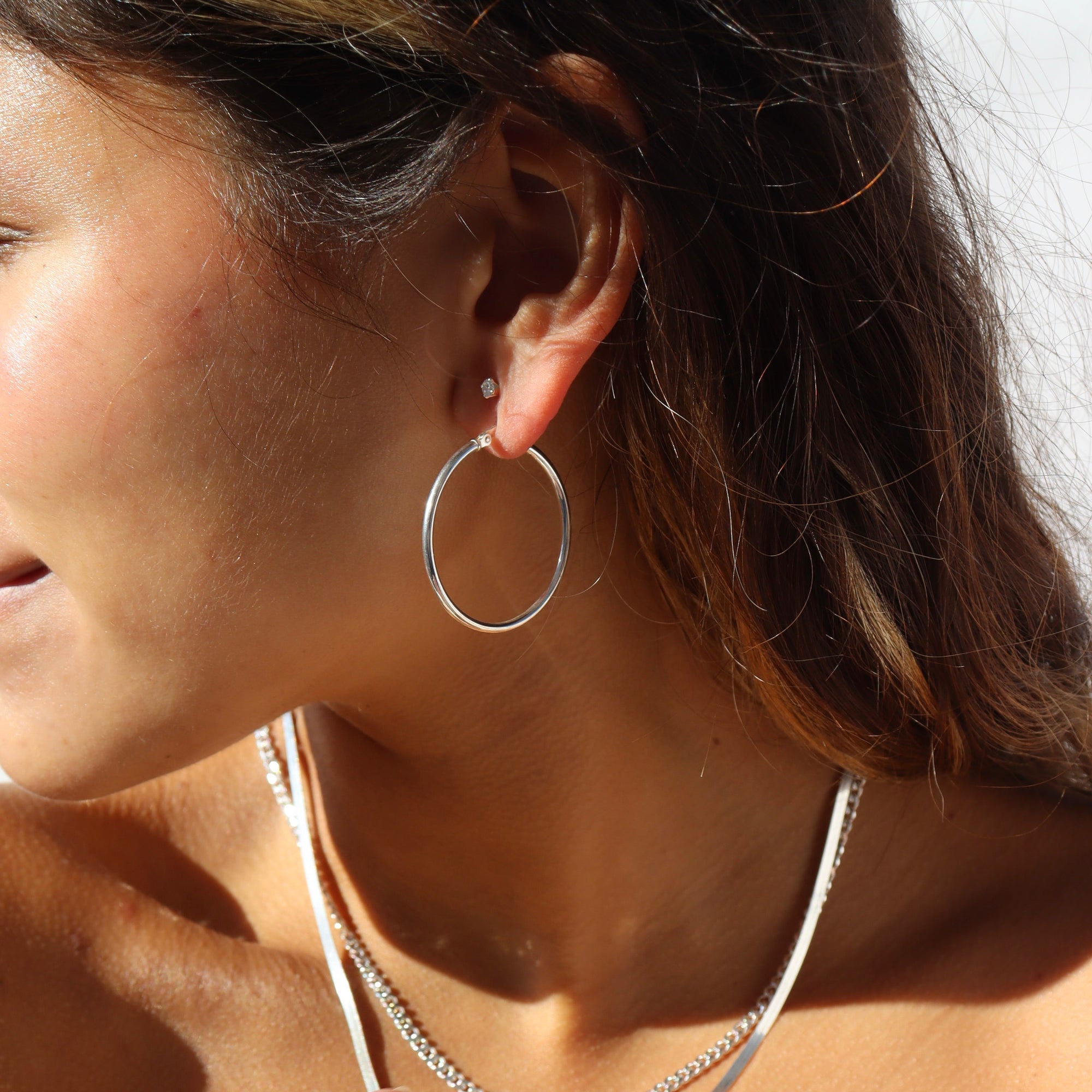 Close-up side profile of a woman with dark hair in bright sunlight, wearing classic large-sized silver hoop earrings, a tiny silver stud, and a stack of silver chain necklaces.