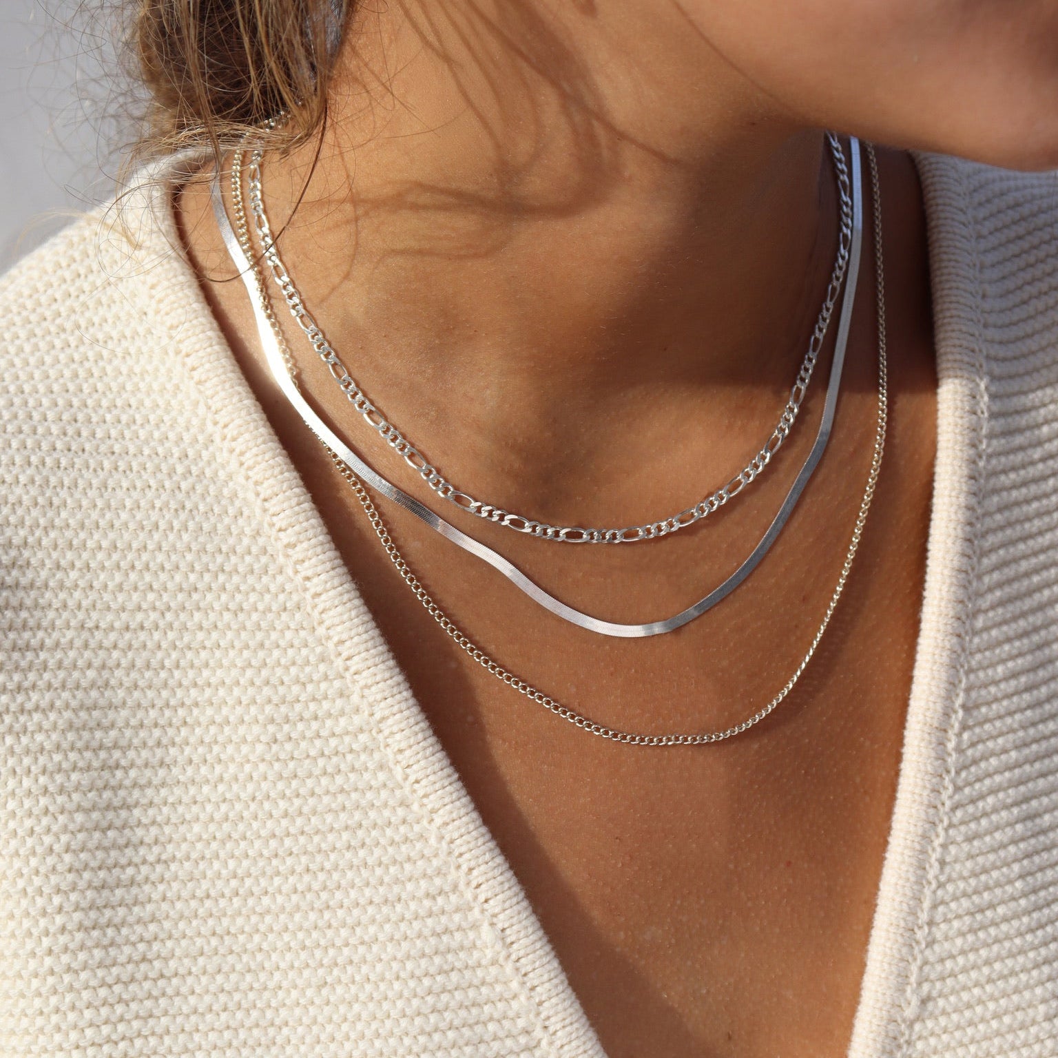 Close-up shot of a woman's neckline in warm natural sunlight, styling a three-tier sterling silver chain set: a top Figaro chain, a sleek middle herringbone snake chain, and a delicate bottom curb chain paired with a cream knit sweater.