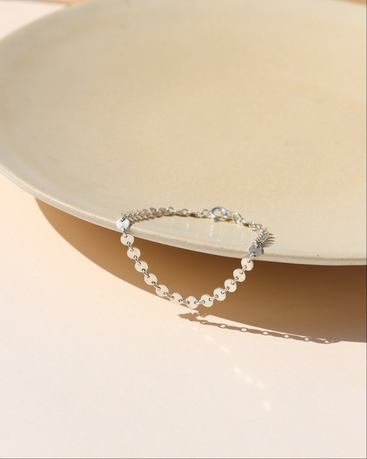 A delicate silver chain bracelet made of small flat disc links, draped over the edge of a cream ceramic platter on a warm beige background.