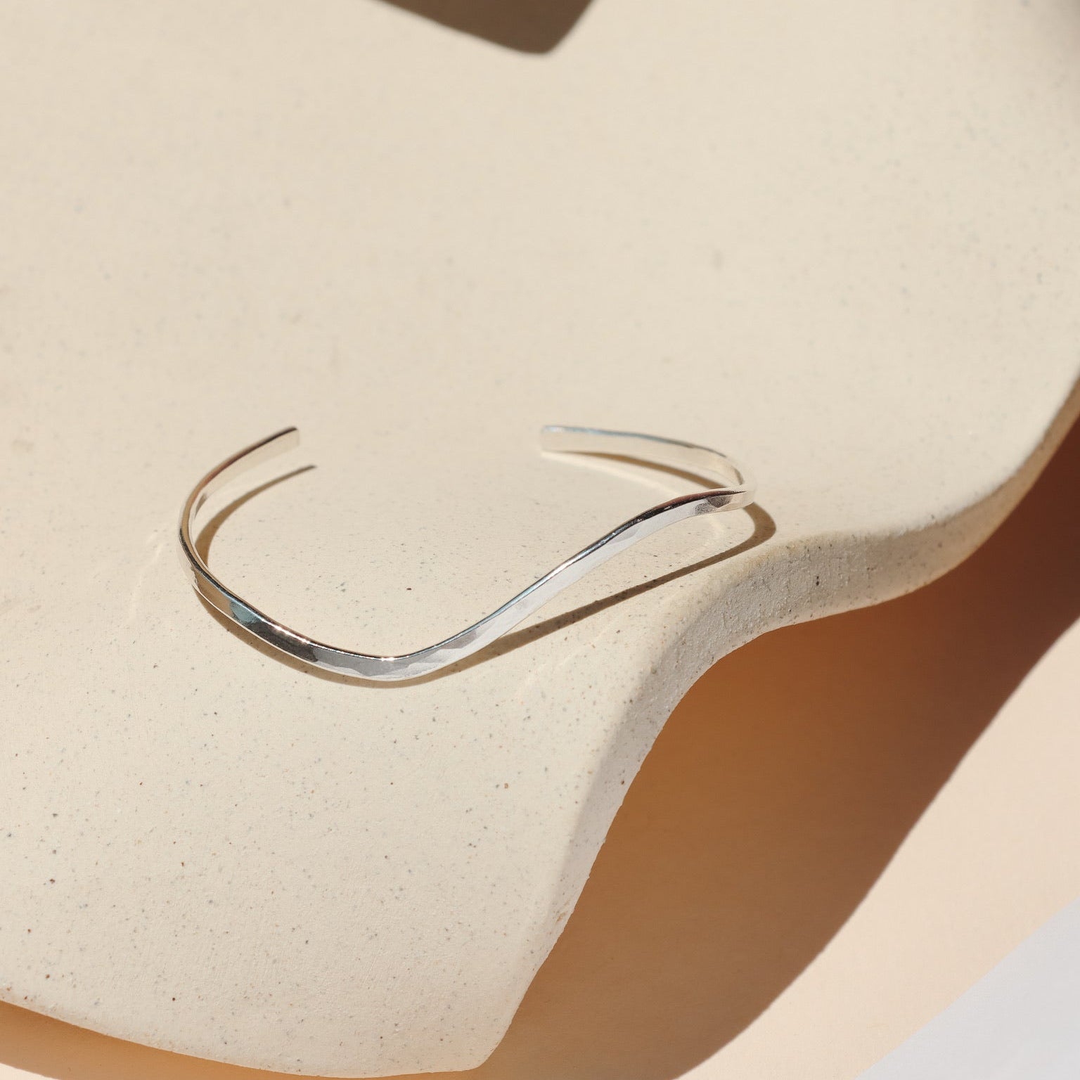 Minimalist sterling silver open cuff bracelet featuring a subtle, fluid wave design, resting on a speckled cream ceramic dish under bright natural light.