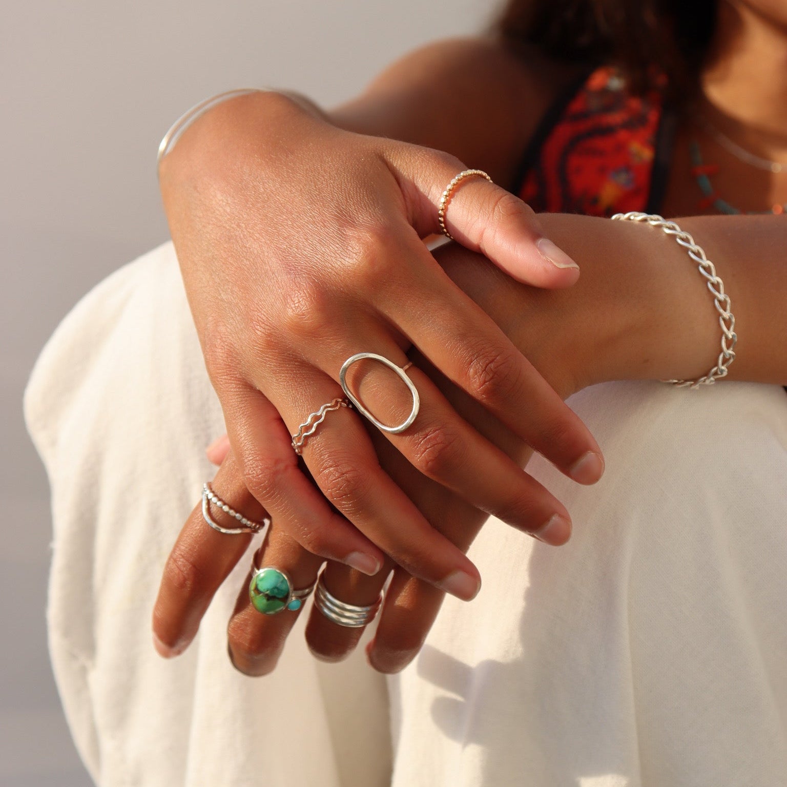 Lifestyle image of layered sterling silver rings, including a bold oval statement ring, natural turquoise ring, beaded stacking rings, and a wavy band paired with a sterling silver chain bracelet in warm natural sunlight.