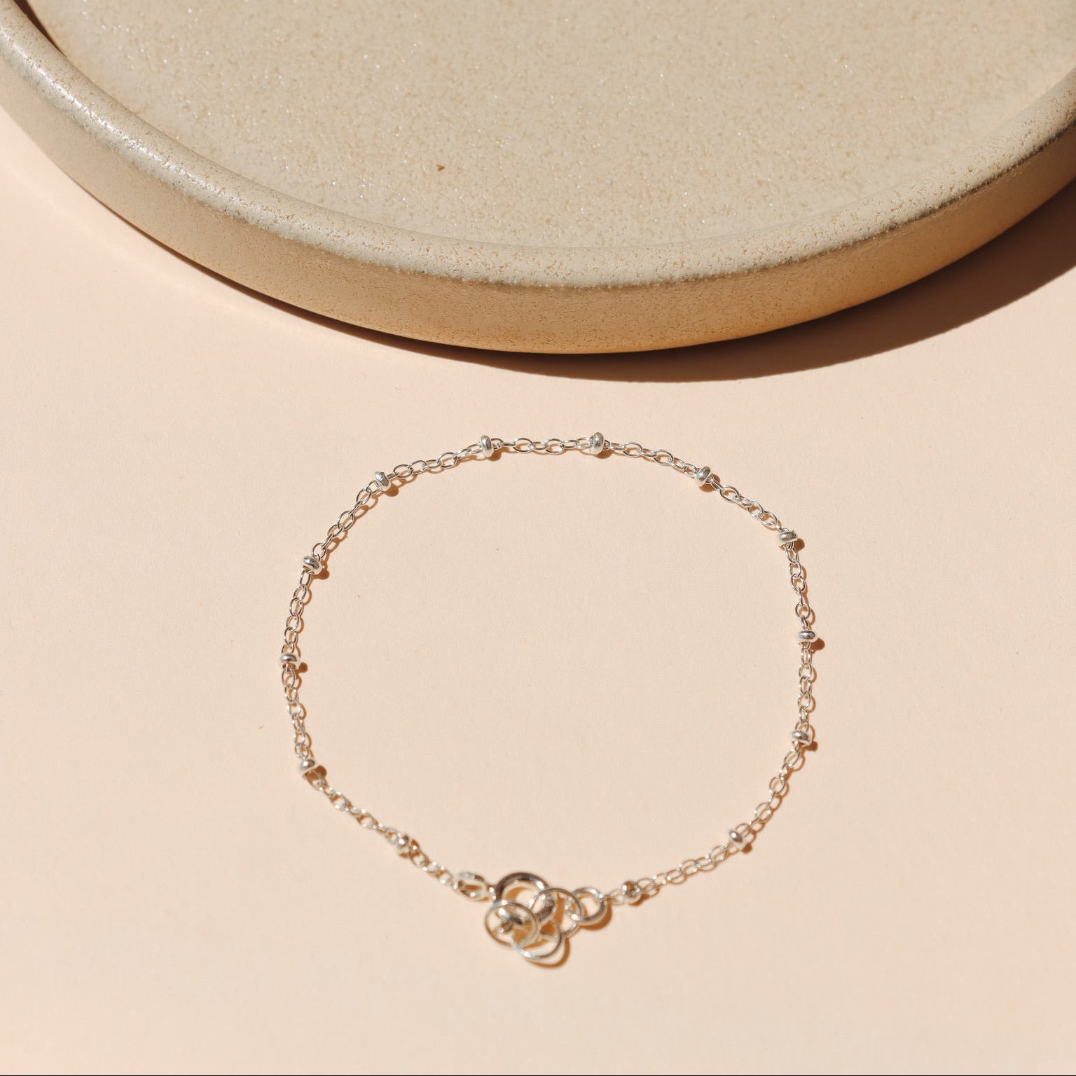 Minimalist fine silver bracelet featuring a thin chain with evenly spaced bead station accents, arranged in a circle on a warm beige background near a smooth ceramic tray.