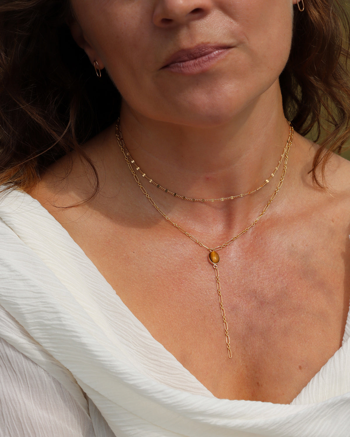 Woman wearing layered gold necklaces with a tiger’s eye lariat necklace and delicate gold chain, styled with a white top.