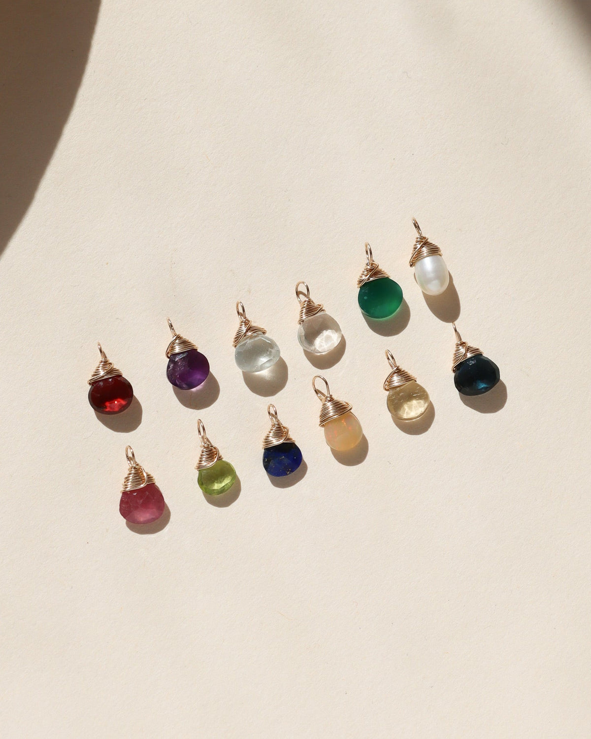 Assortment of colorful faceted gemstone charms in red, purple, pink, green, blue, opal, and pearl tones, each finished with gold wire wrapping, arranged on a cream background in warm sunlight.