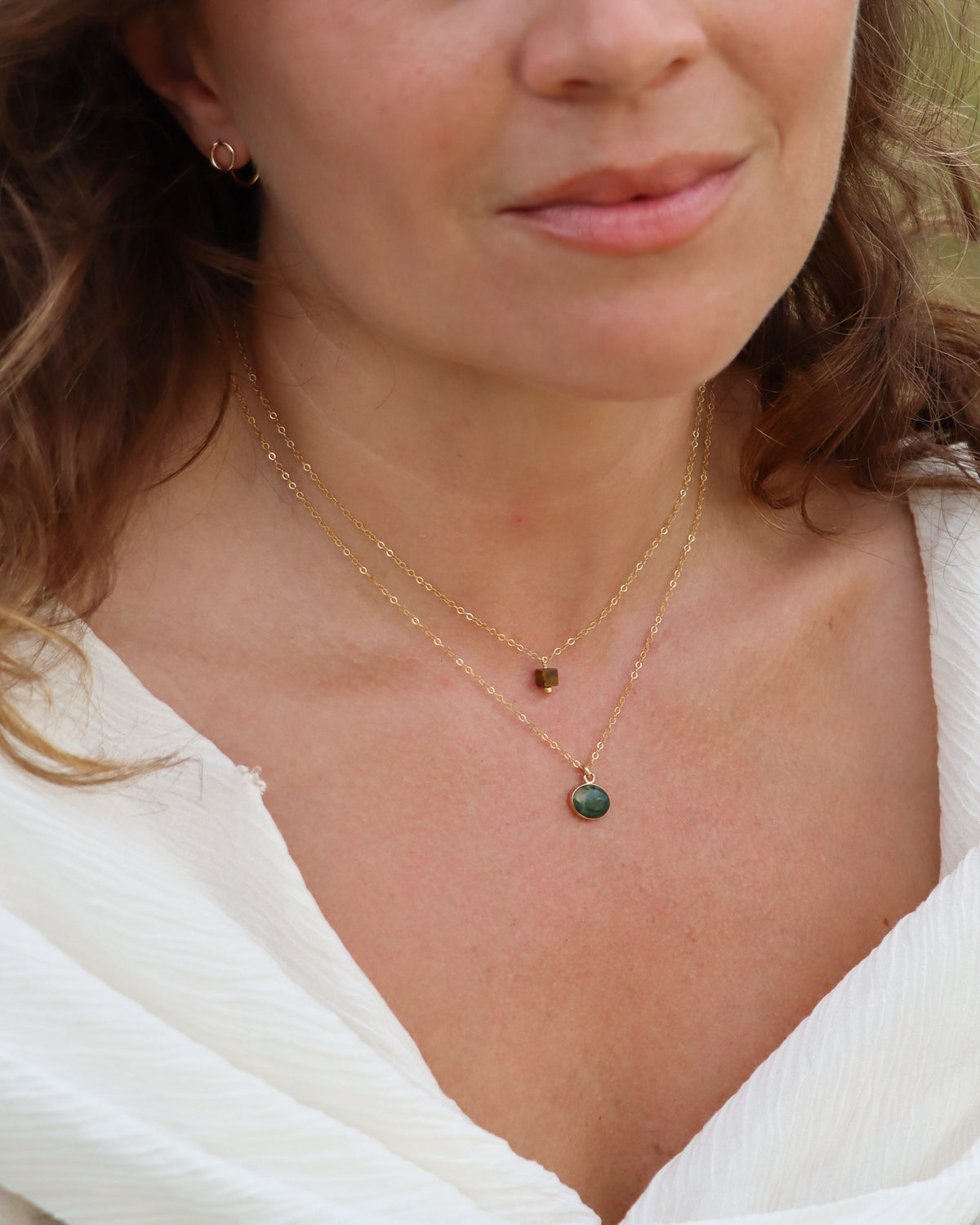 Layered gold necklaces featuring a small green gemstone pendant and a brown bead pendant, styled with a white blouse.