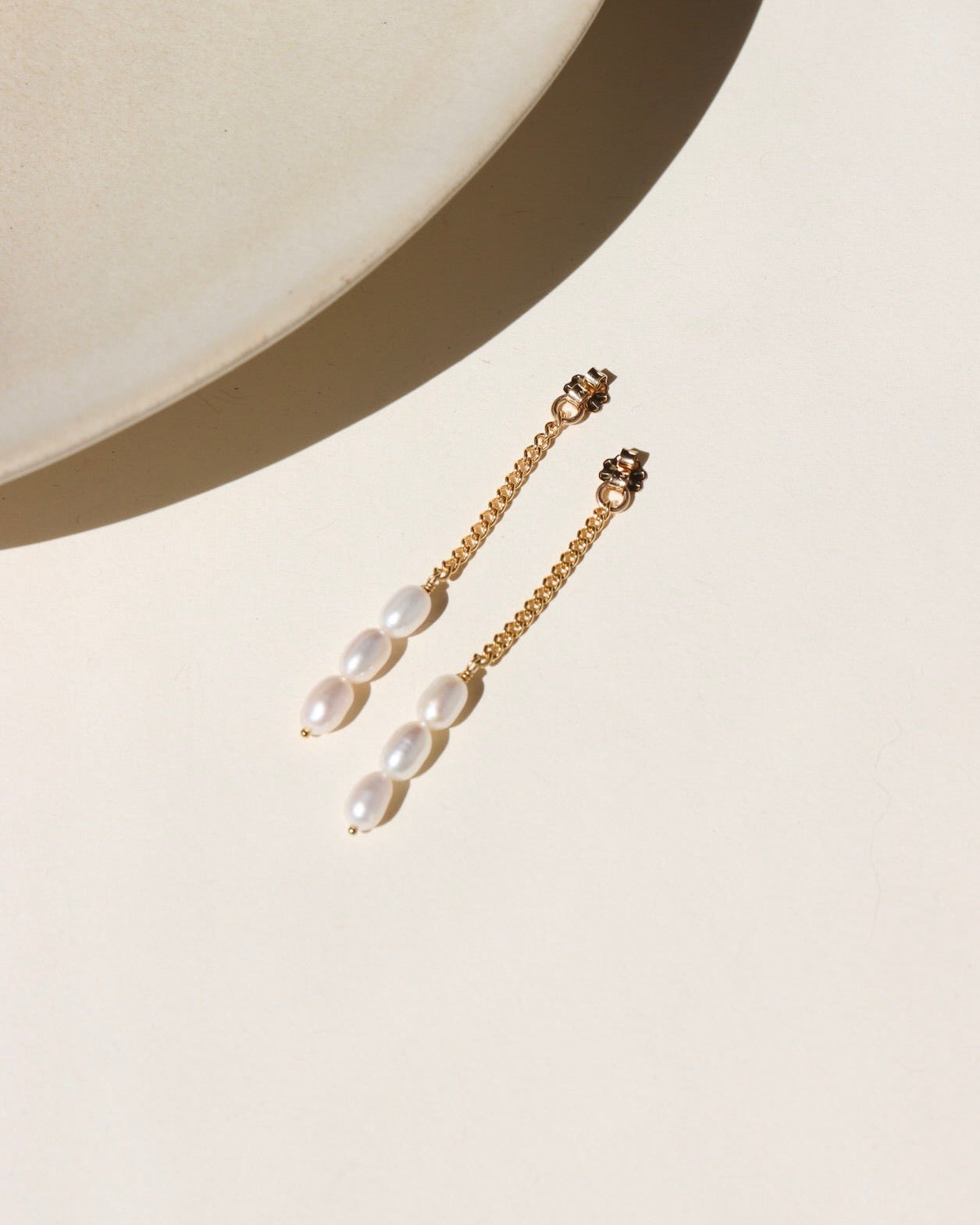 A 14k gold fill curb chain backdrop with three trillium pearls at the bottom of each chain, the backdrops are displayed on a cream display in the sunlight 