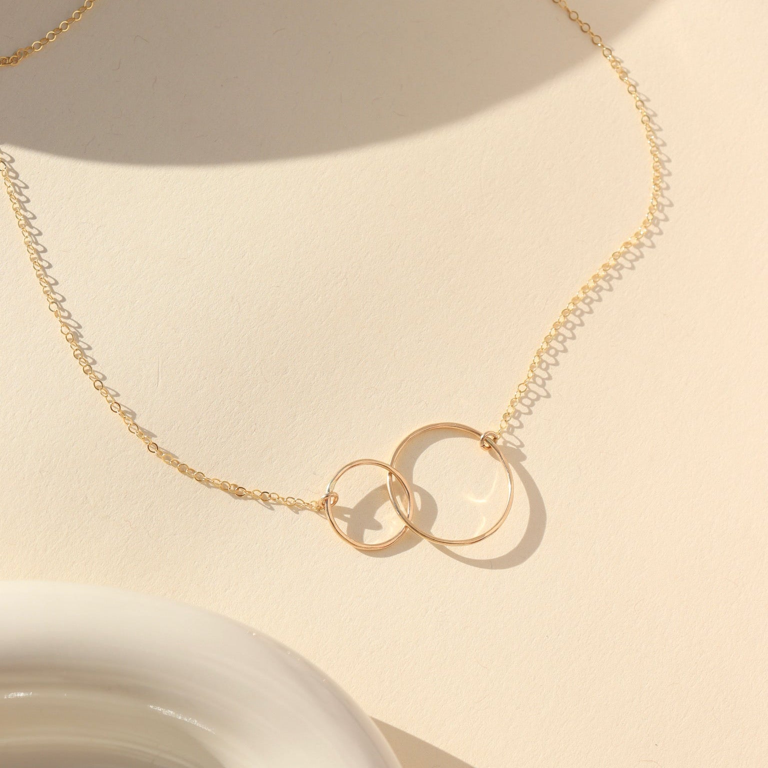 Top-down view of a fine gold chain necklace with an interlocking double-circle pendant, laid out on a cream-colored surface with warm natural sunlight casting subtle shadows next to a white ceramic dish