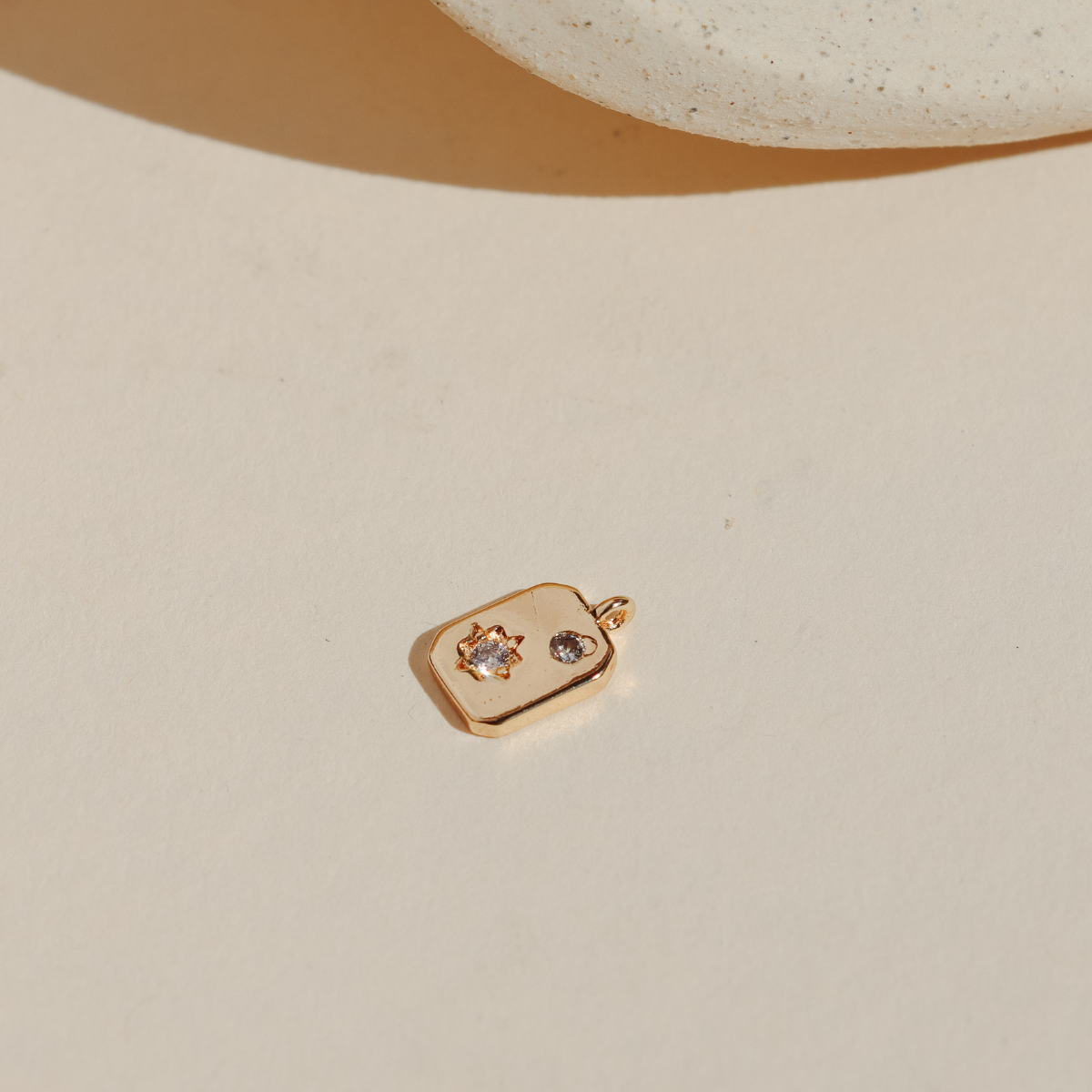 Mini gold celestial charm featuring a sparkling sun and moon design on a rectangular tag, photographed on a neutral surface in warm natural light.