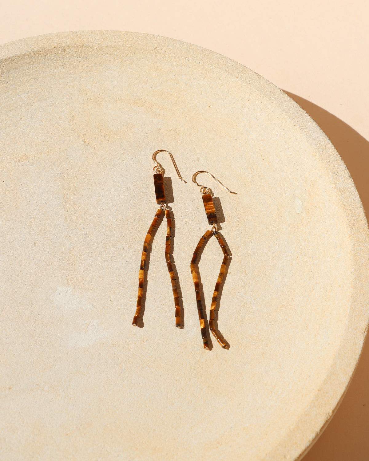 A pair of dangling earrings with gold filled french ear wires and rectangular Tiger's Eye gemstones and two long, Tiger's Eye beaded strands dangling, displayed on a textured beige ceramic dish.
