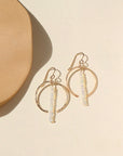 A 14k gold fill hoop featuring white opal gemstones dropped 2" from earring post. Earrings are displayed on a cream back drop in the sunlight. 