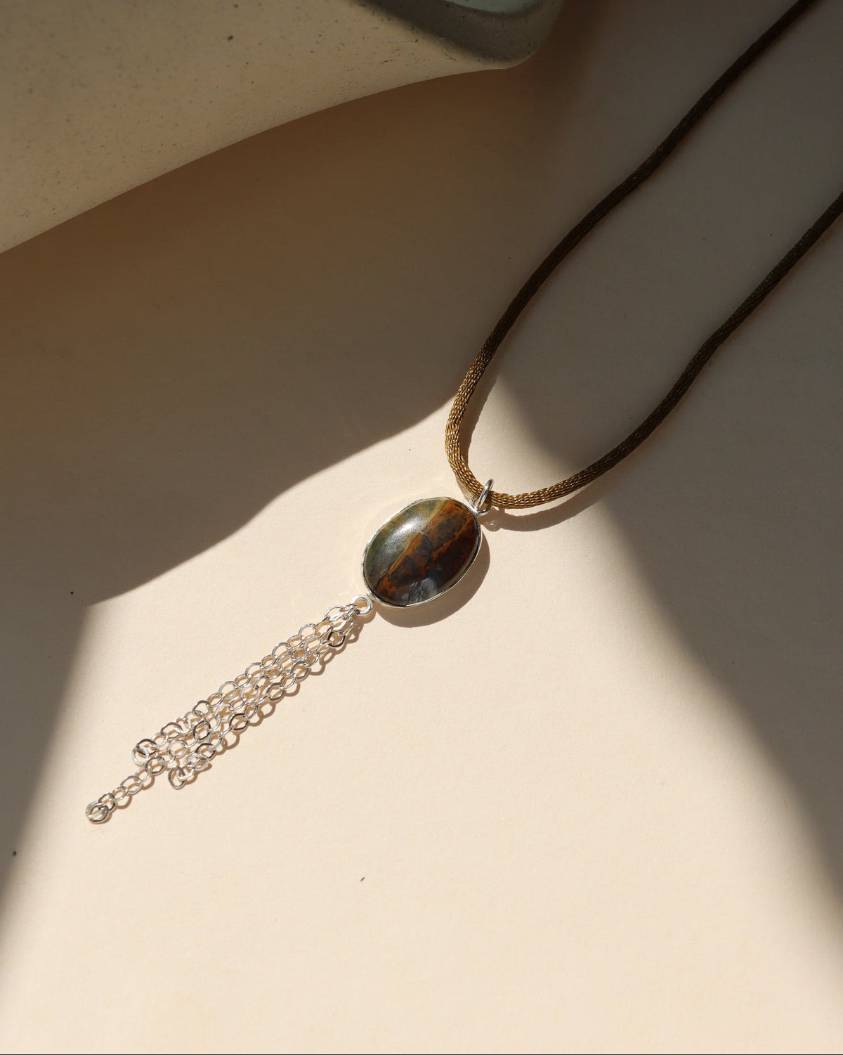 Oval brown and gray gemstone pendant necklace with silver chain fringe, displayed in warm sunlight on a neutral background.