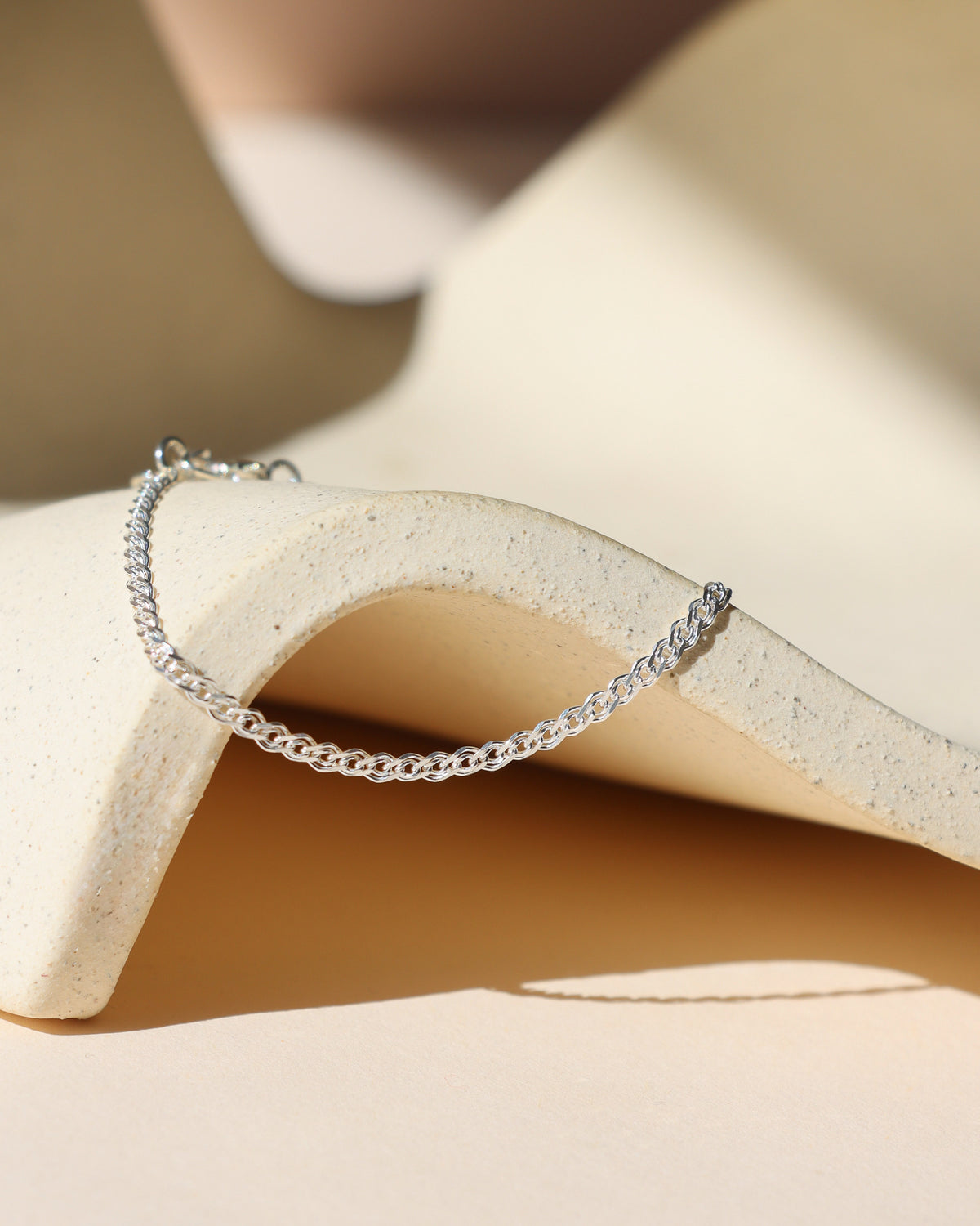 Delicate sterling silver woven chain bracelet displayed on a sculptural neutral surface in warm sunlight.