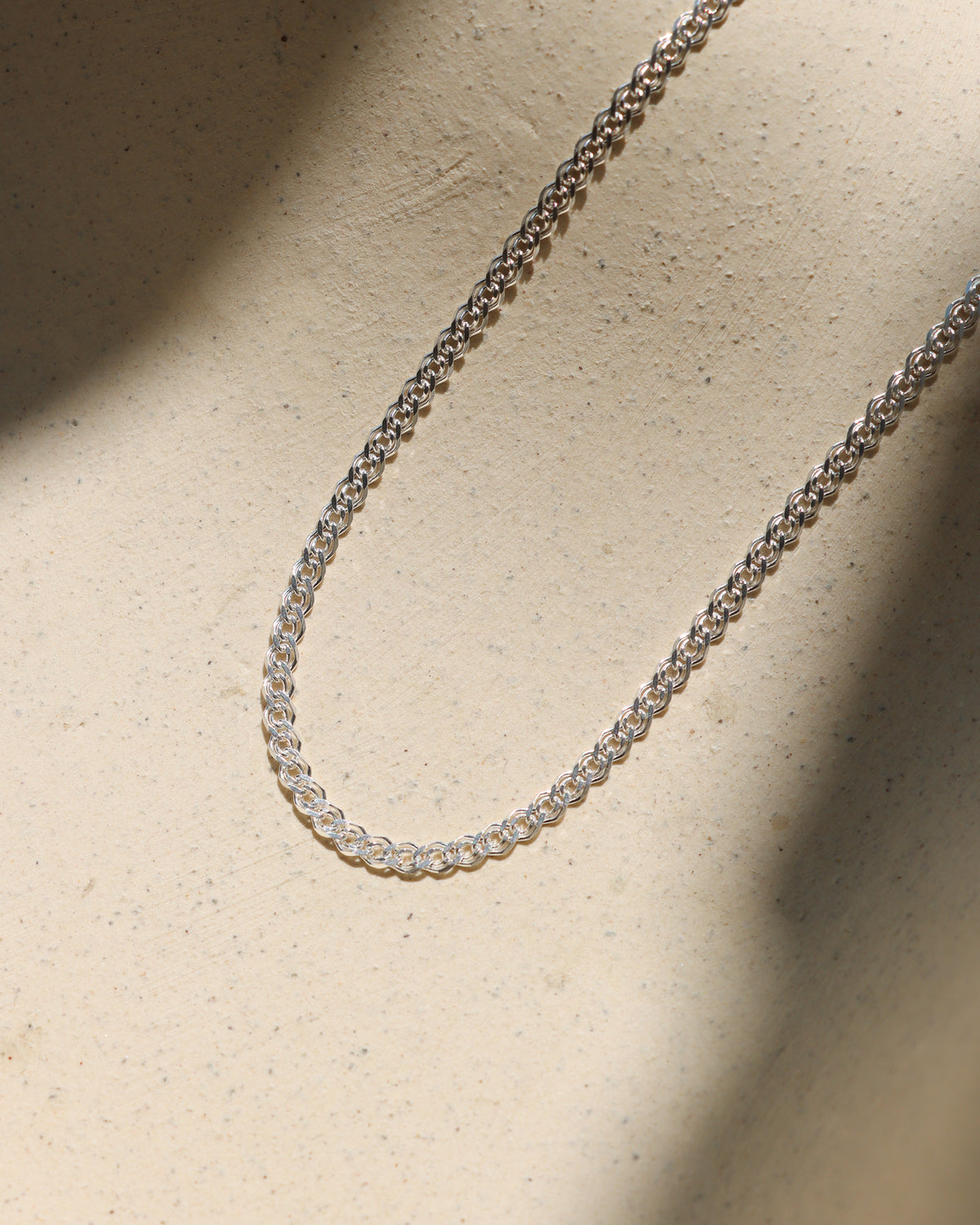 Sterling silver woven chain necklace displayed on a neutral textured surface in warm sunlight.