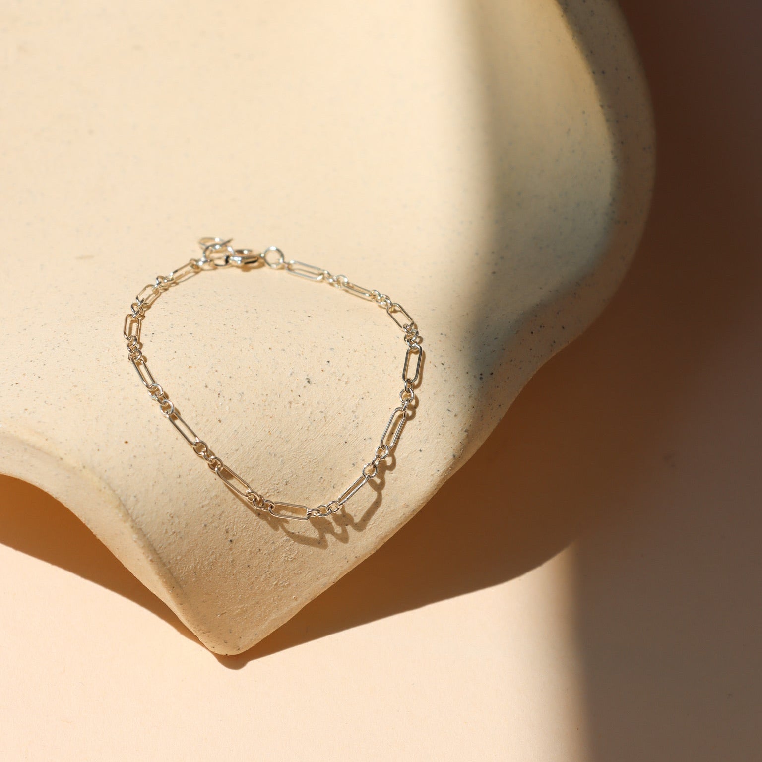 Minimalist fine silver paperclip link chain bracelet laid out on a beige speckled ceramic prop, bathed in soft, warm sunlight with subtle shadows.