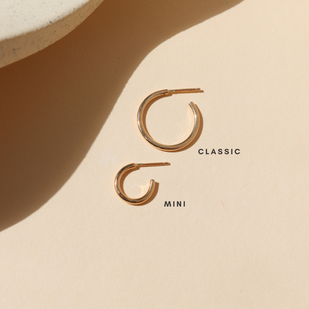 Two polished gold open hoop earrings in classic and mini sizes, displayed on a warm cream background with natural sunlight and soft shadows to show the difference in size.